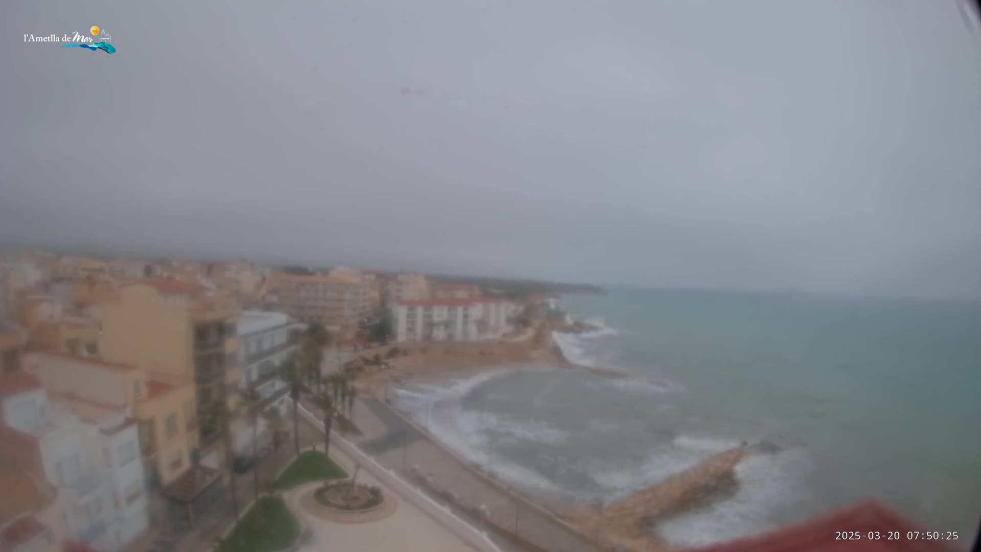A hazy, overcast day reveals a coastal town with buildings along a shoreline where moderate waves crash against a rocky breakwater.