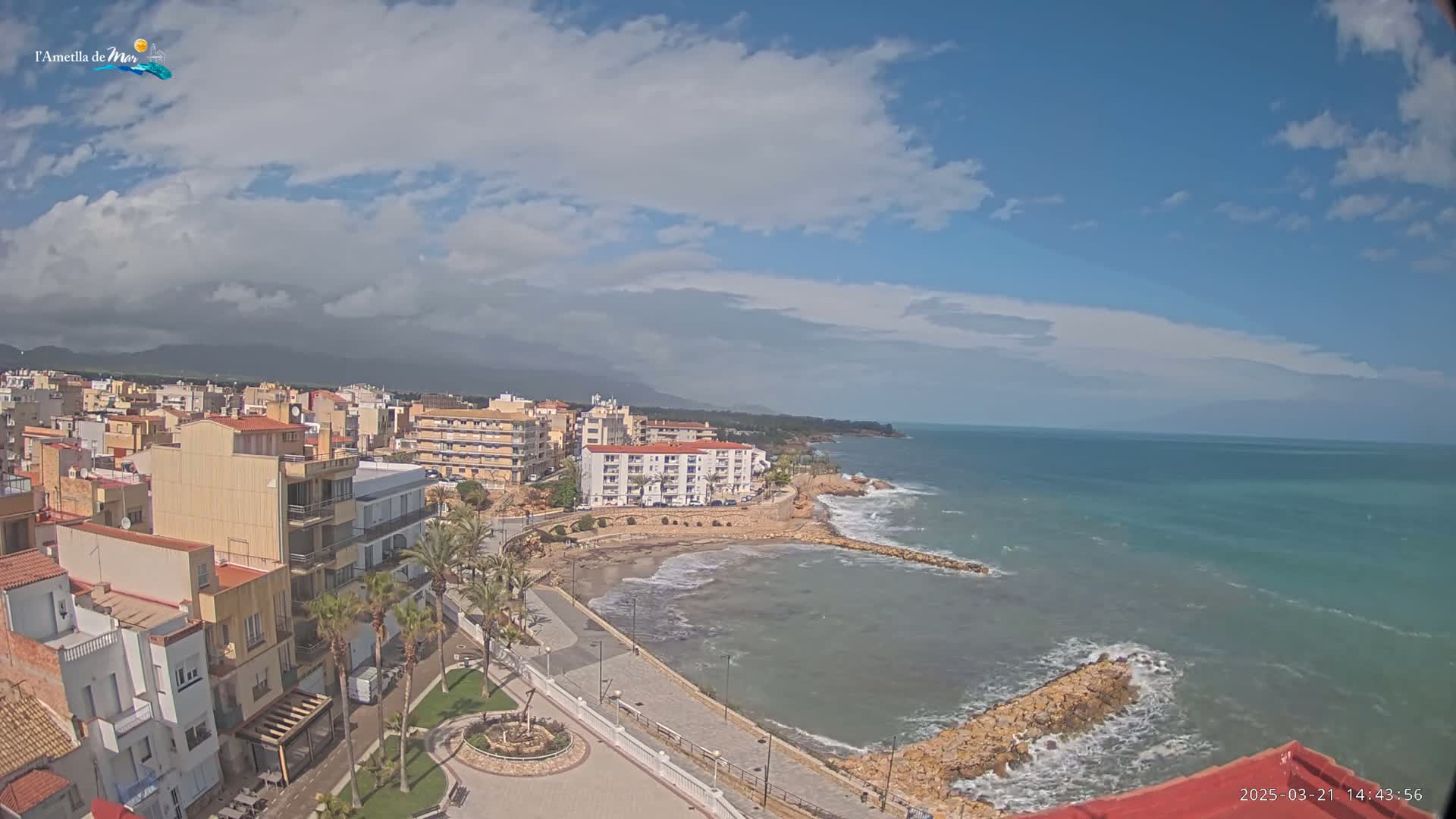 An elevated view shows a coastal town under a partly cloudy sky, with a calm sea lapping against a rocky shoreline.