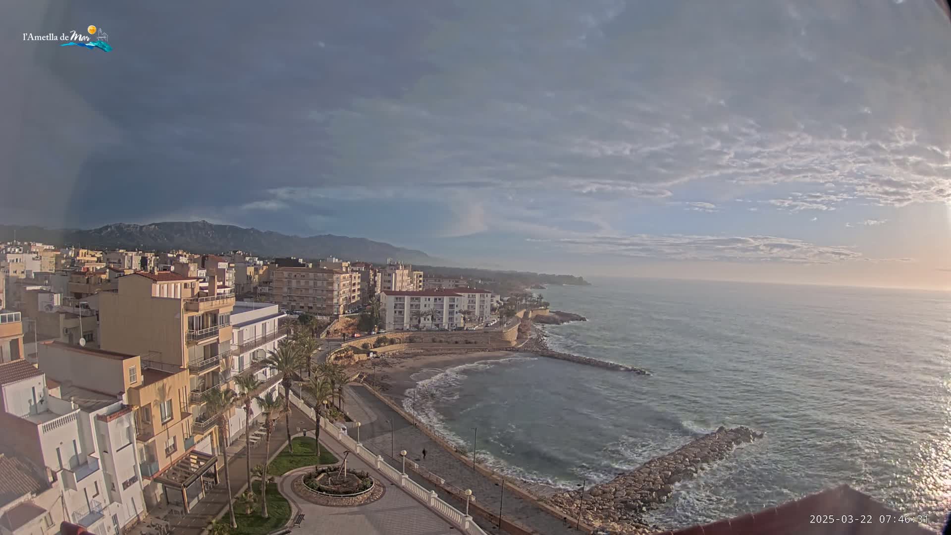 A coastal town with buildings, palm trees, and a paved walkway along a calm sea under a partly cloudy sky.