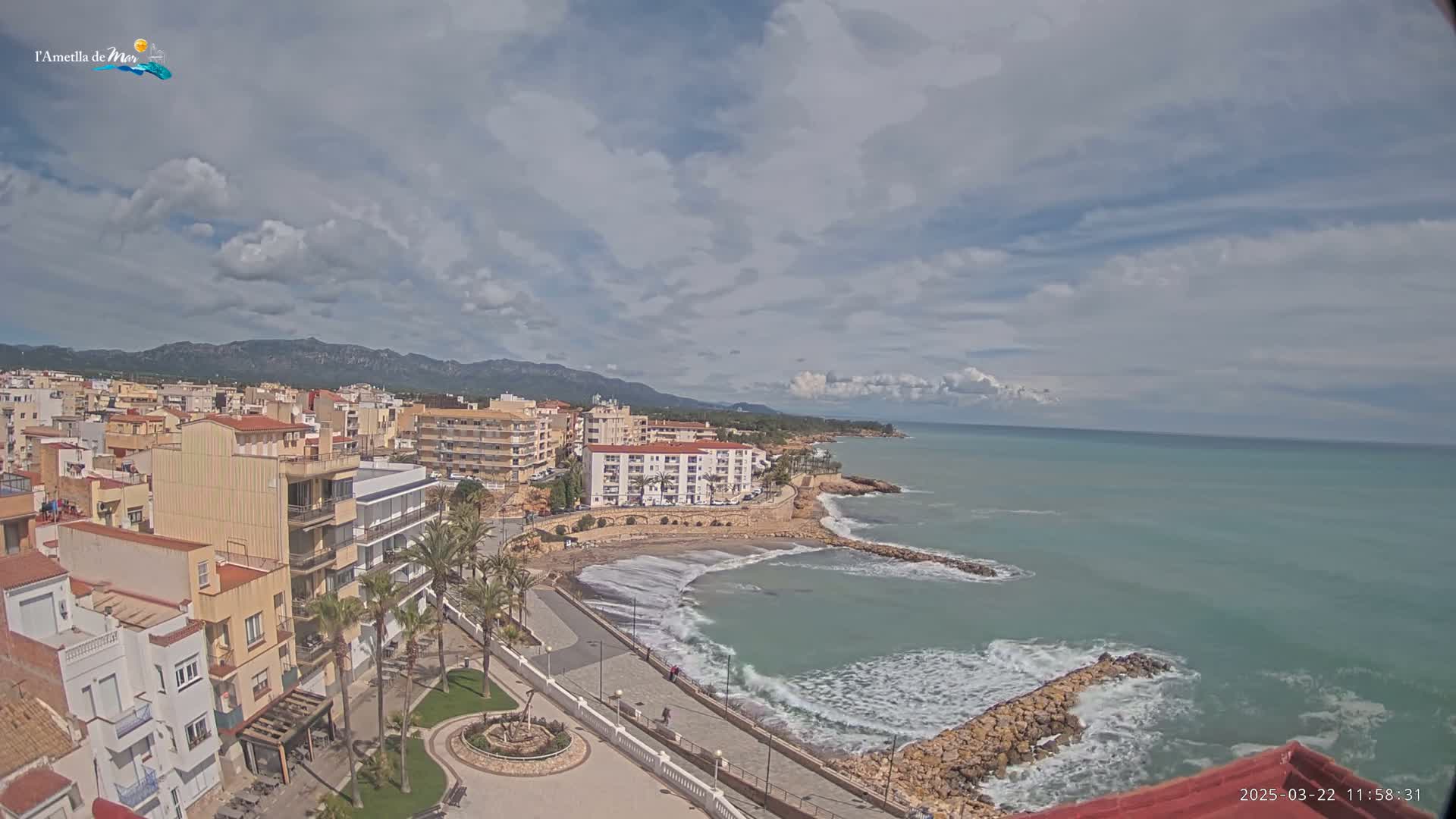 A coastal town with buildings, palm trees, and a promenade overlooks a partially cloudy sky and a sea with small waves breaking against a rock wall.
