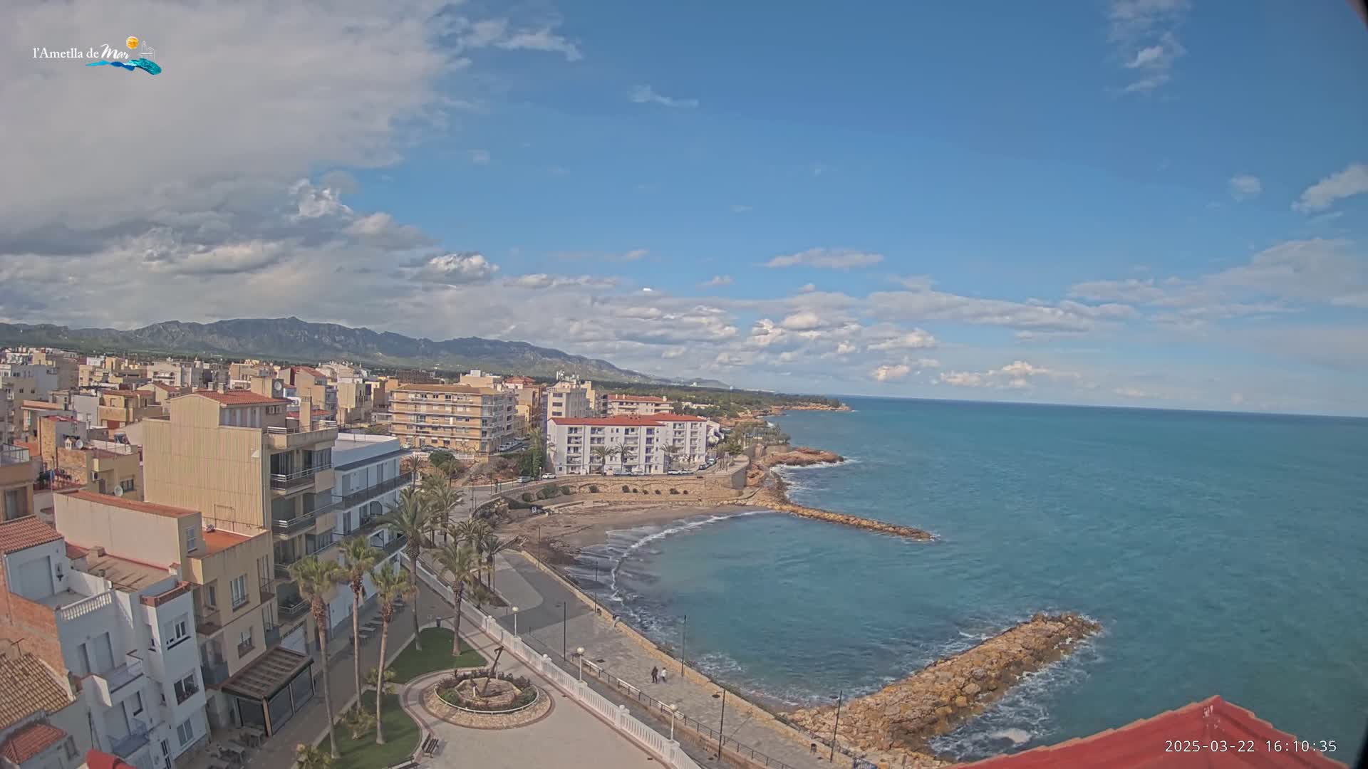 A coastal town with buildings and palm trees sits on a partly cloudy day next to a calm, teal ocean with small waves, mountains visible in the distance.