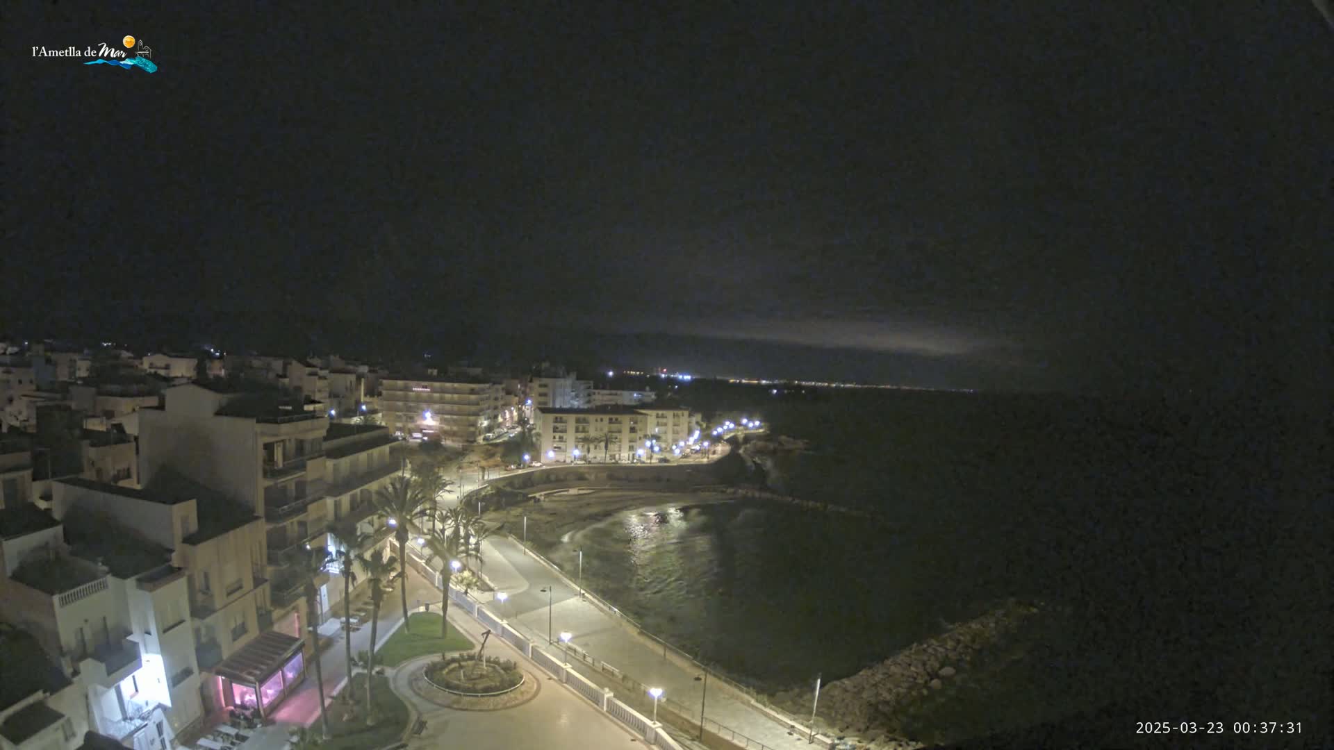 A nighttime coastal town scene shows a dark, calm sea next to a paved walkway lined with palm trees and buildings with lights on.