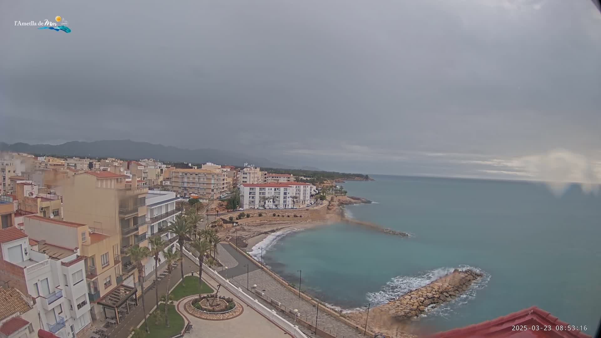 A coastal town under an overcast sky features buildings along a shoreline with a calm, teal sea and a rocky breakwater.