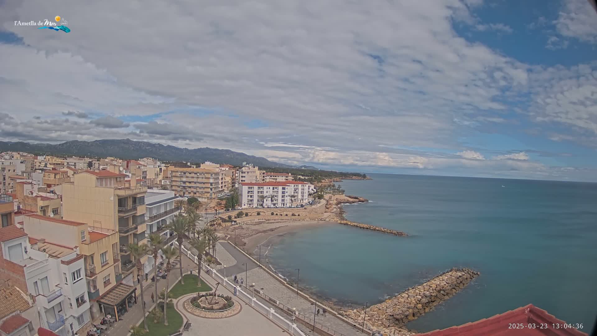 A partly cloudy day reveals a coastal town with buildings, a small beach, and a calm sea under a mostly cloudy sky, with mountains visible in the distance.