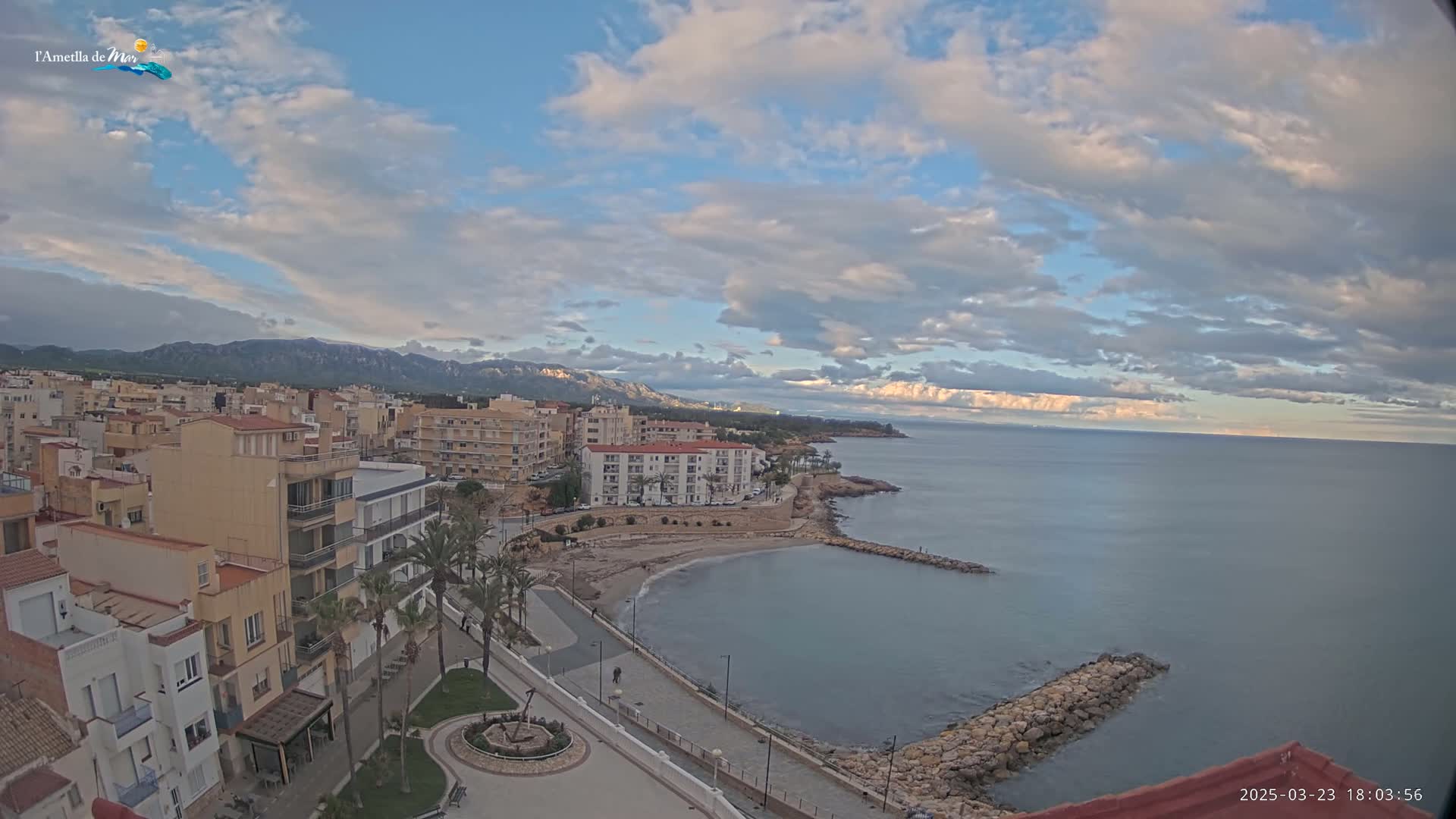 A coastal town with buildings, palm trees, and a small beach is under a partly cloudy sky, with mountains visible in the distance.