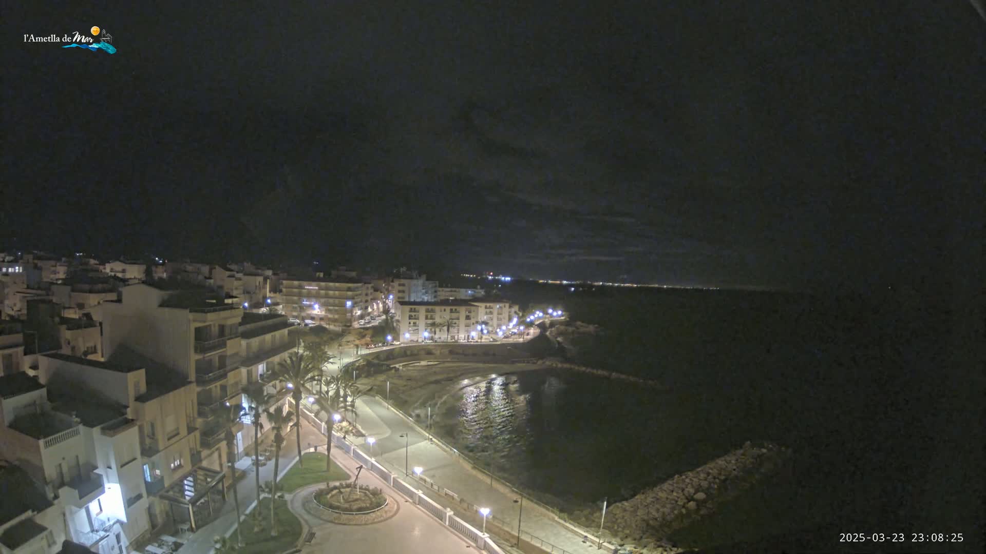 A nighttime, aerial view of a coastal town with buildings illuminated along a paved waterfront path next to a dark body of water under a mostly cloudy sky.