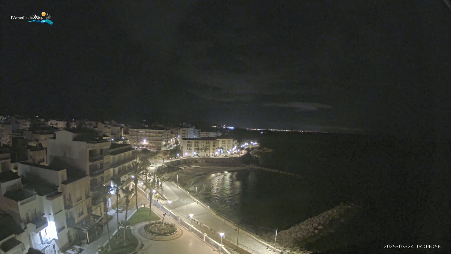 A nighttime, coastal town scene shows a crescent-shaped bay with a paved walkway lined with palm trees and buildings illuminated by streetlights under a dark, mostly cloudy sky.