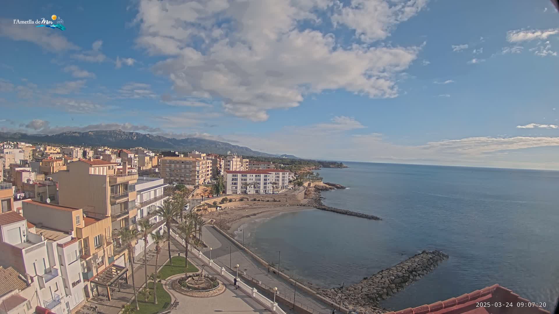 A partly cloudy day shows a coastal town with buildings, palm trees, a small beach, and a calm sea with mountains in the background.