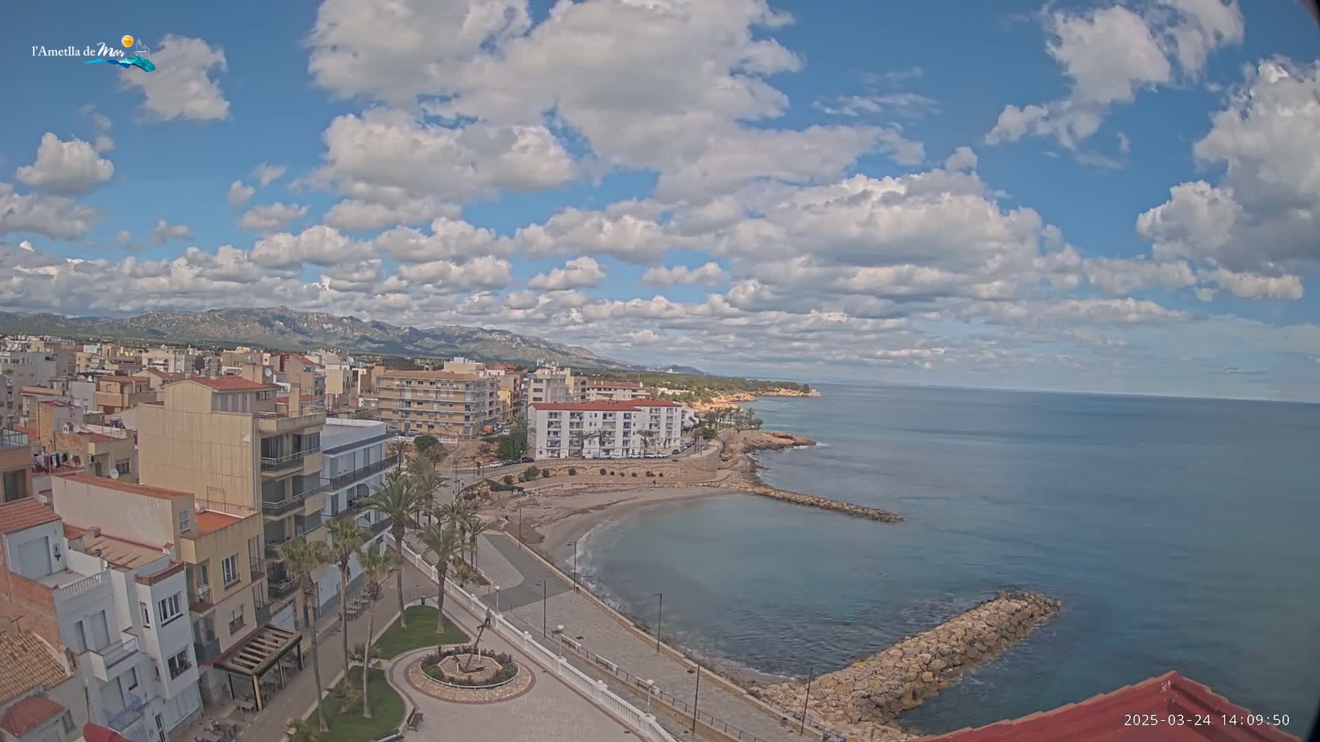 A coastal town with buildings, palm trees, a small beach, and a calm sea under a partly cloudy sky.