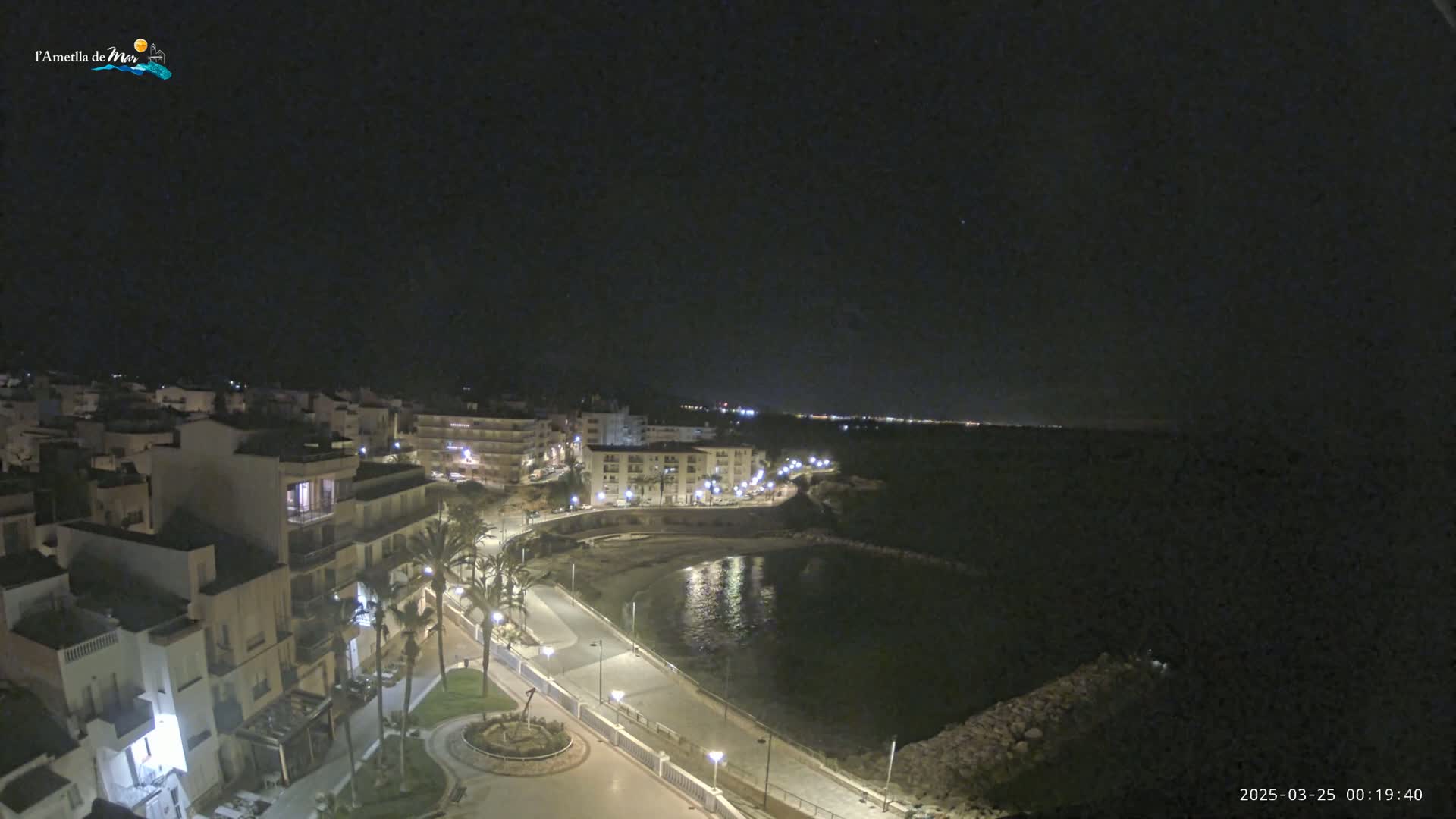 A nighttime aerial view shows a coastal town with buildings illuminated along a paved walkway beside a calm bay under a clear, dark sky.
