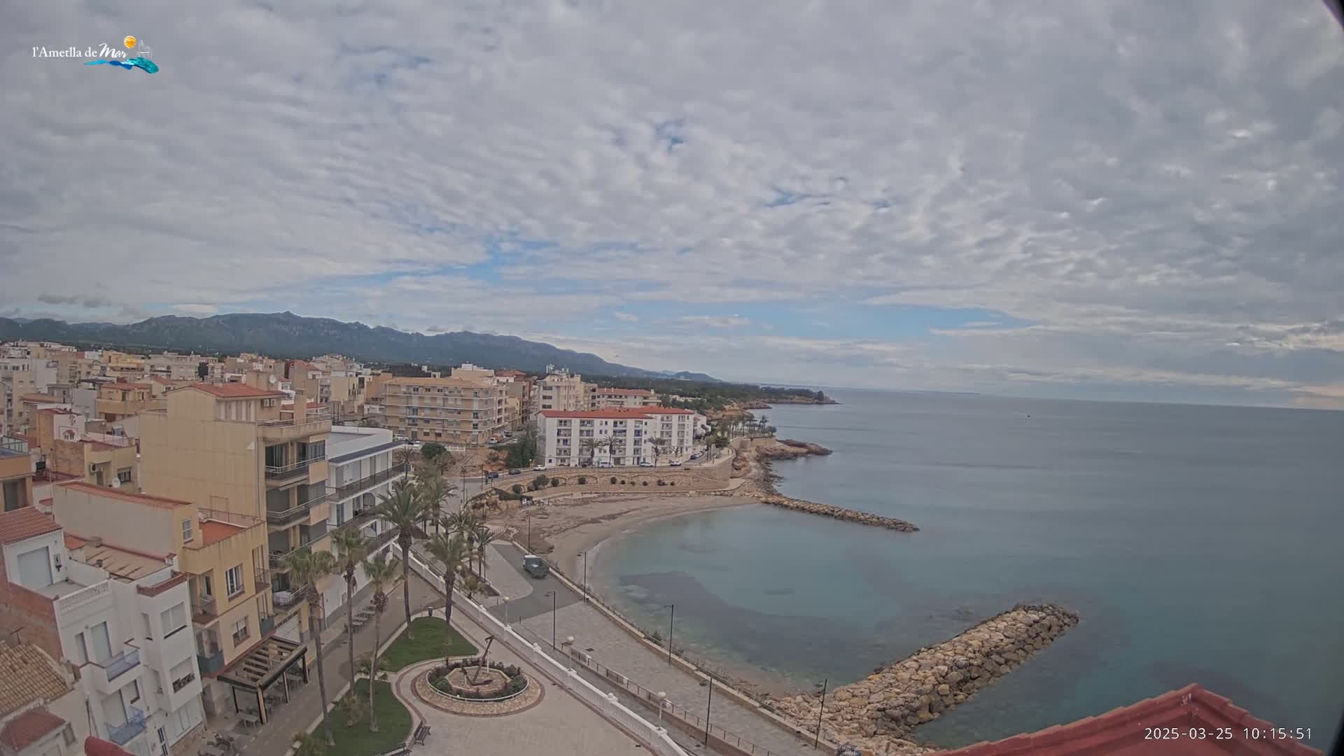 A coastal town with low-rise buildings and palm trees, under a partly cloudy sky, overlooks a calm bay and distant mountains.