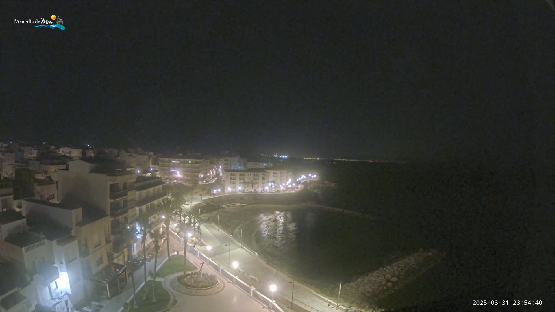 A nighttime, high-angle view shows a coastal town with buildings lit up along a paved walkway next to a dark body of water.