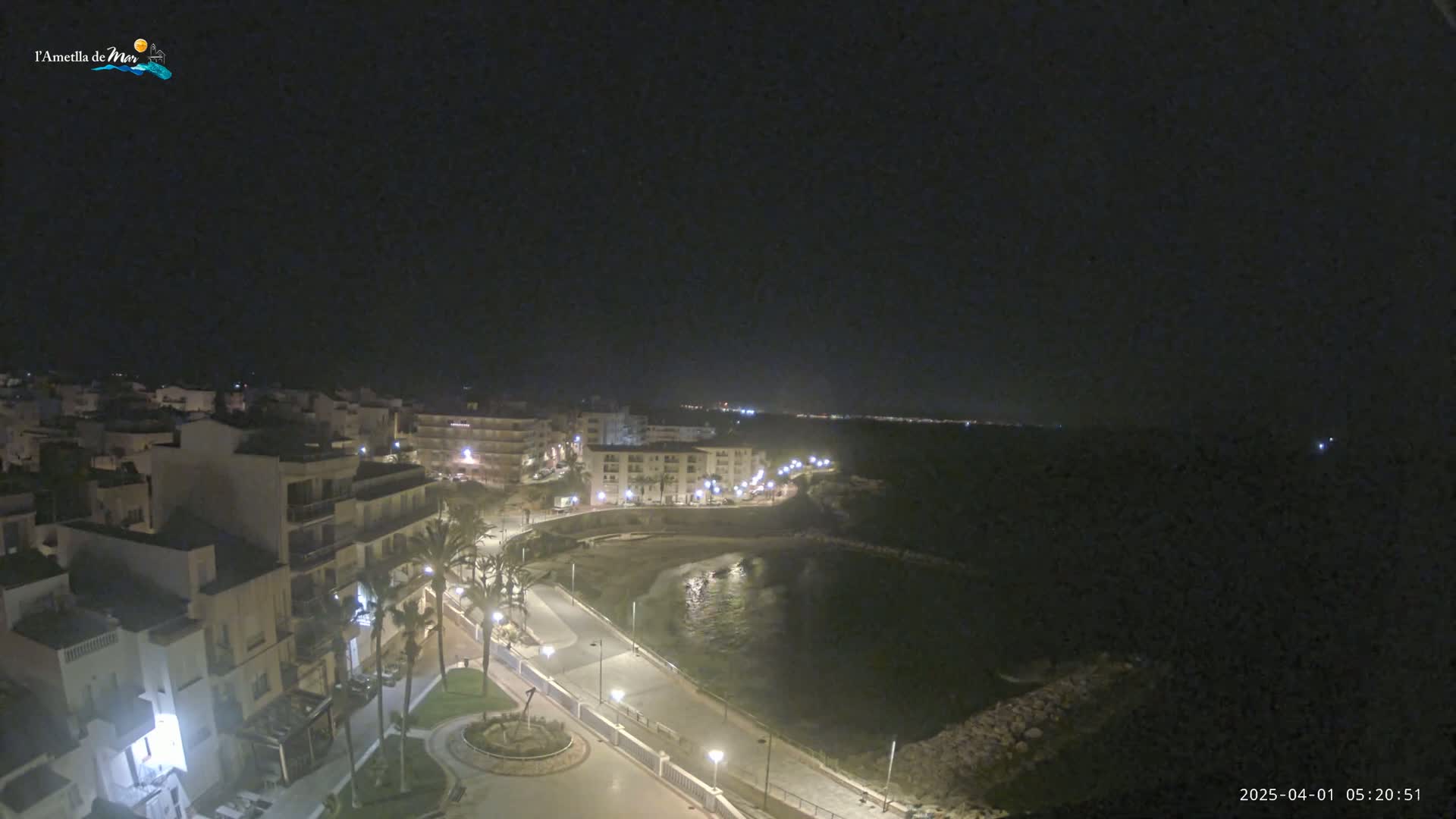 A nighttime, aerial view of a coastal town with buildings illuminated along a paved waterfront path next to a dark body of water.