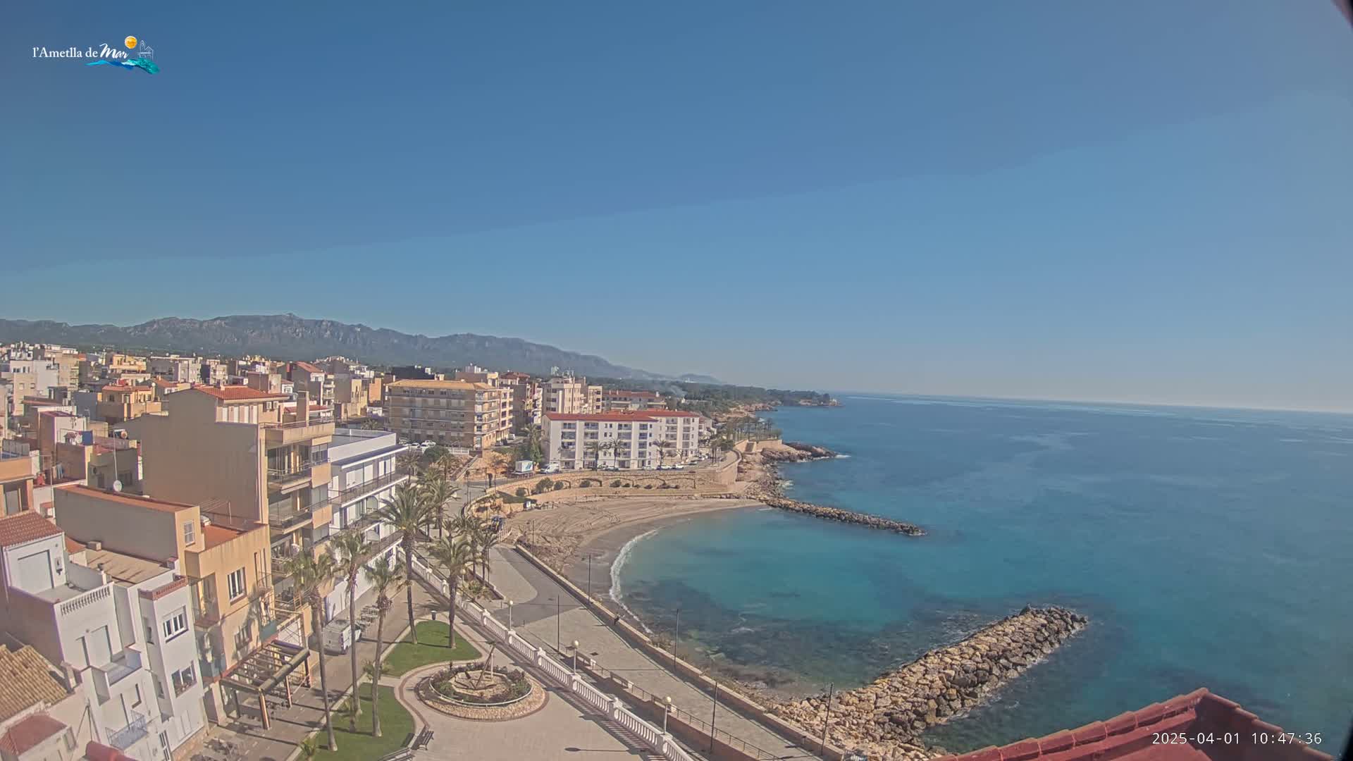 A coastal town with buildings, palm trees, and a small sandy beach is seen on a clear, sunny day, with calm blue water extending to distant mountains.