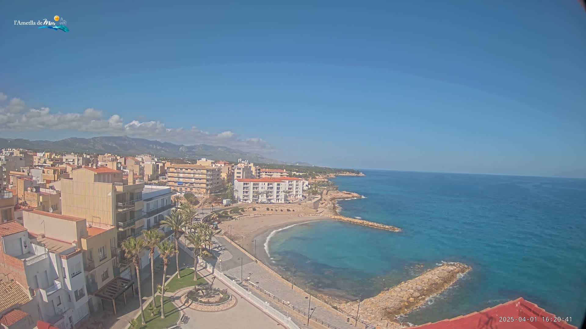 A coastal town with buildings, palm trees, and a small sandy beach is nestled against a calm, blue sea under a mostly clear, sunny sky.