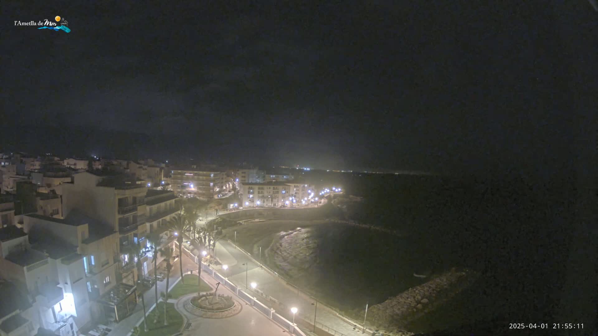 A nighttime, high-angle view of a coastal town with illuminated buildings and streets curving along a dark body of water under a clear, dark sky.
