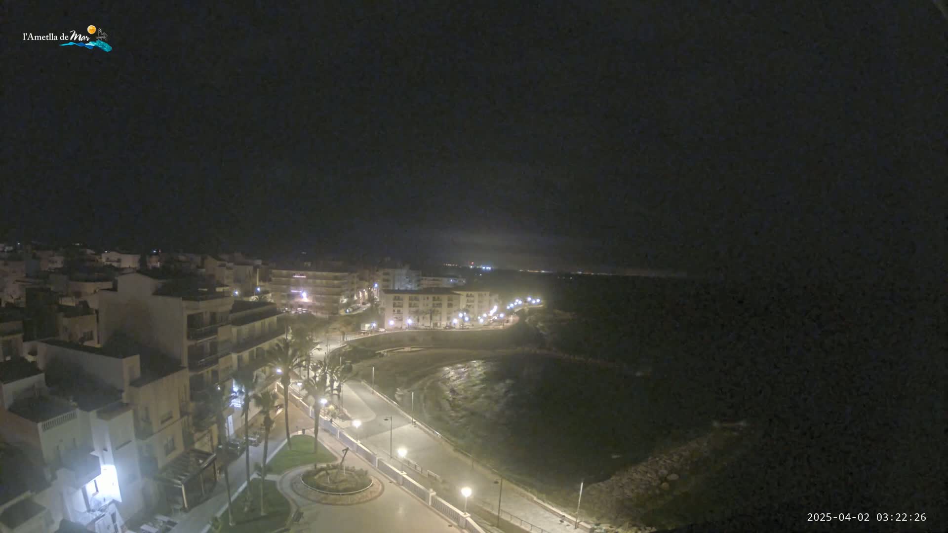 An aerial nighttime view shows a coastal town with buildings illuminated along a curved road that leads to a dark, calm body of water.
