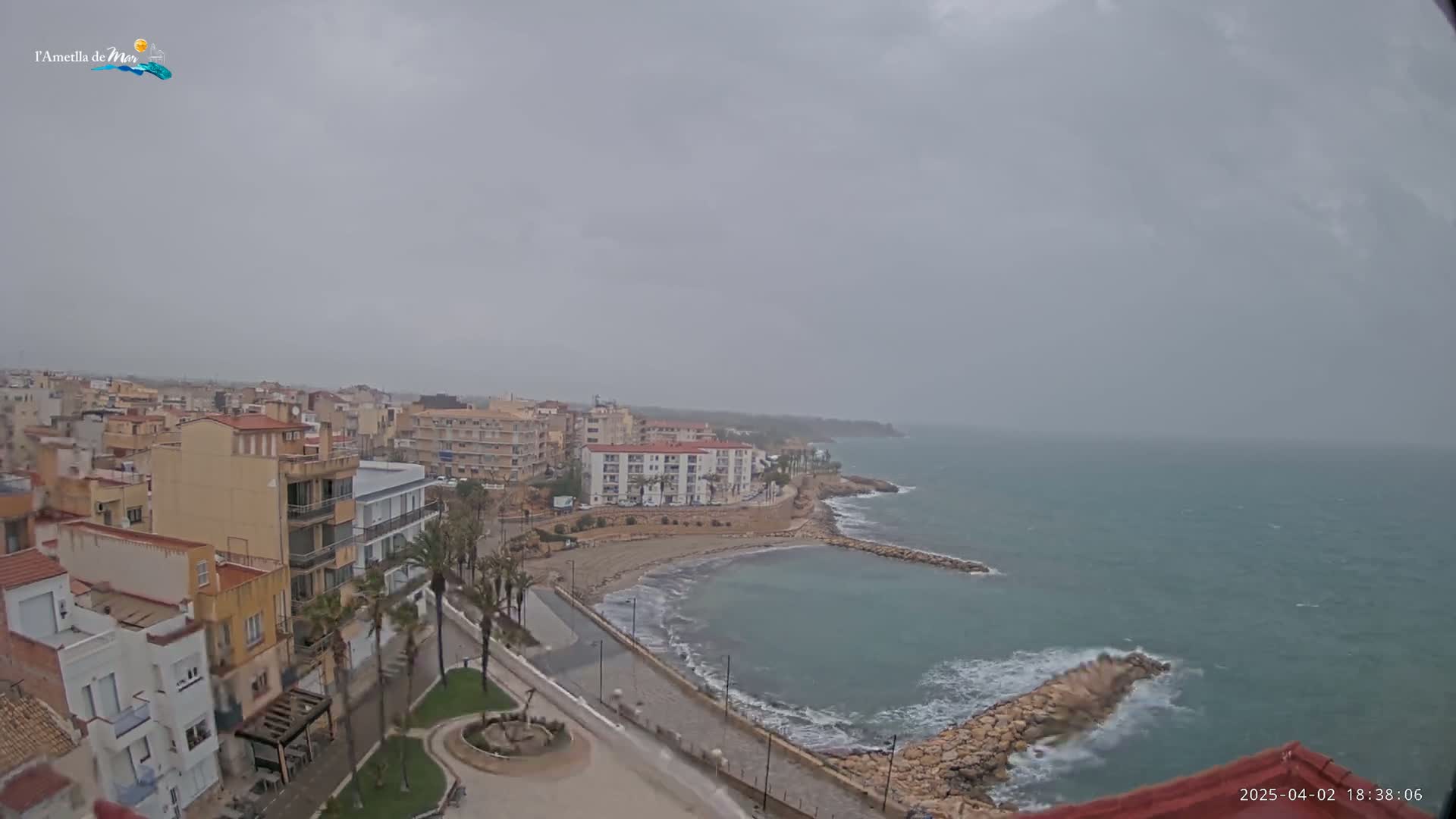 A coastal town with low-rise buildings is seen under an overcast sky, with a somewhat choppy sea visible beyond a small bay and a rocky breakwater.