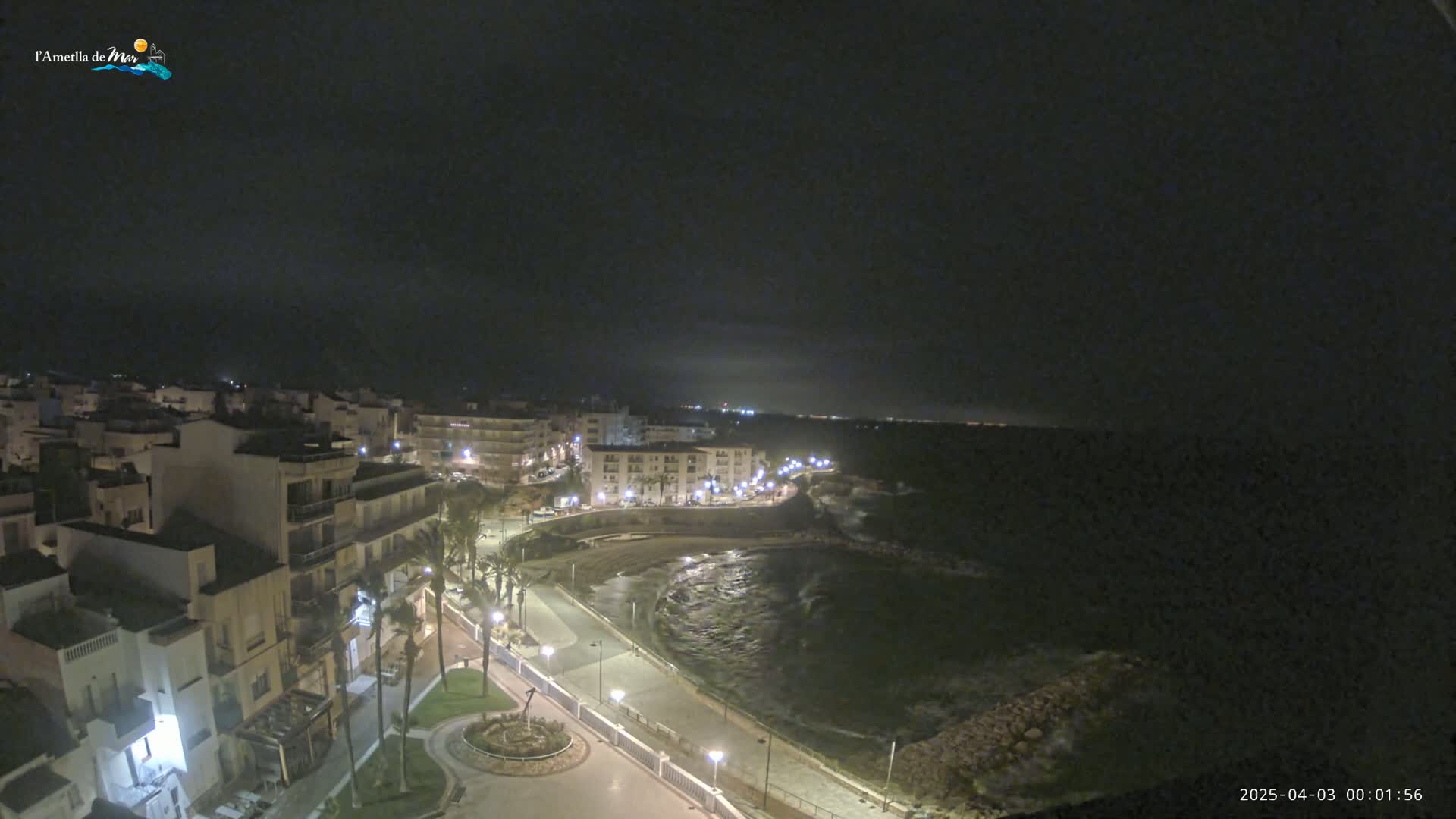 A nighttime, coastal town scene shows a dark, wavy ocean under a mostly clear sky, with a paved walkway and buildings lit up along the shore.