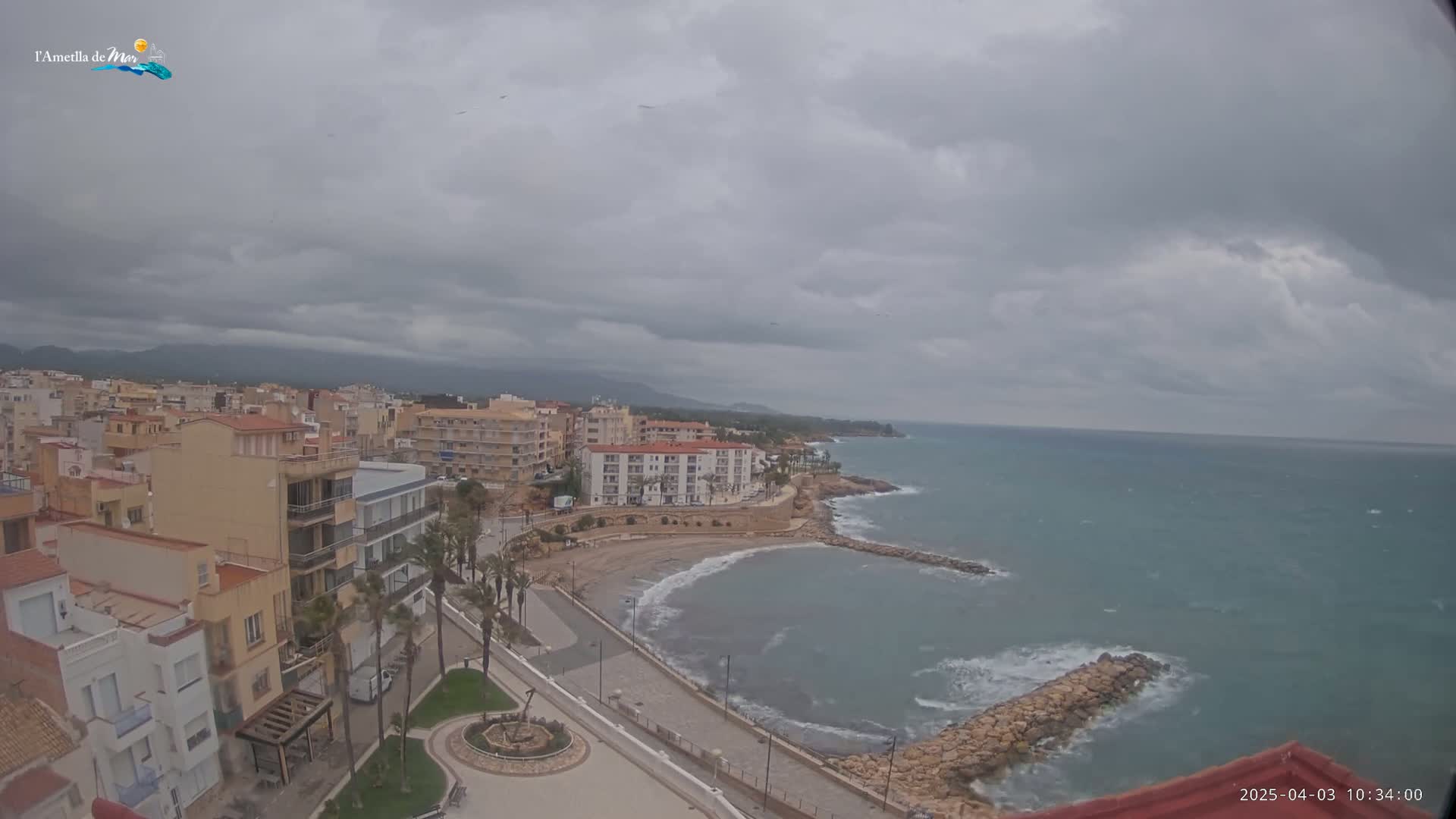 An overcast day shows a coastal town with multi-story buildings, a small beach, and a rocky breakwater extending into a choppy sea.
