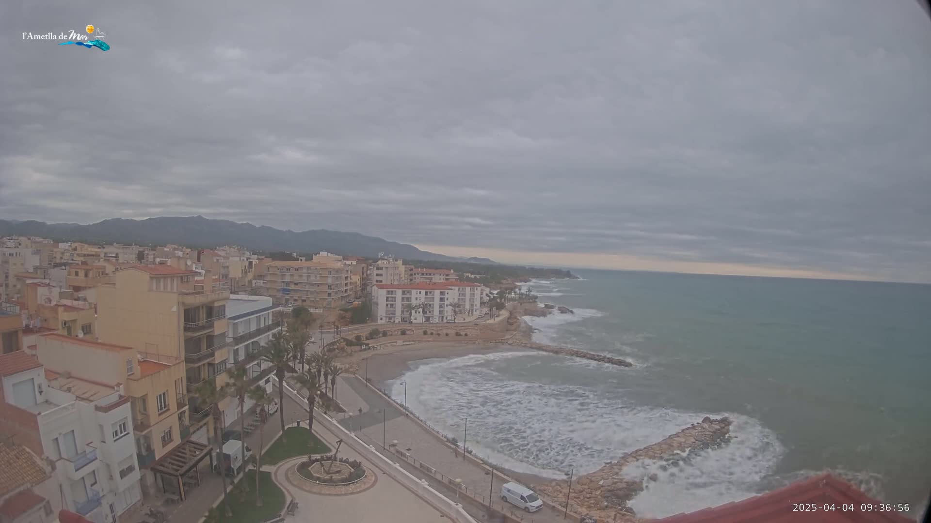 A coastal town with multi-story buildings and palm trees sits under an overcast sky, with a choppy sea and rocky coastline visible in the foreground.