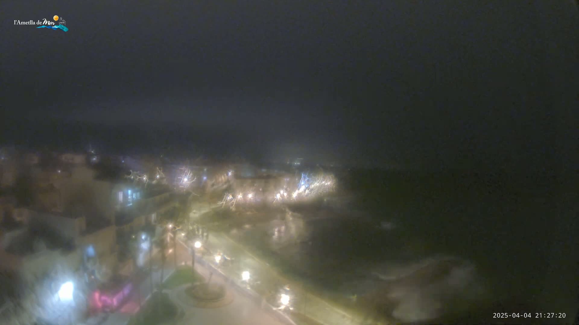 A foggy night scene shows a dimly lit coastal town with streetlights illuminating a walkway along the water.