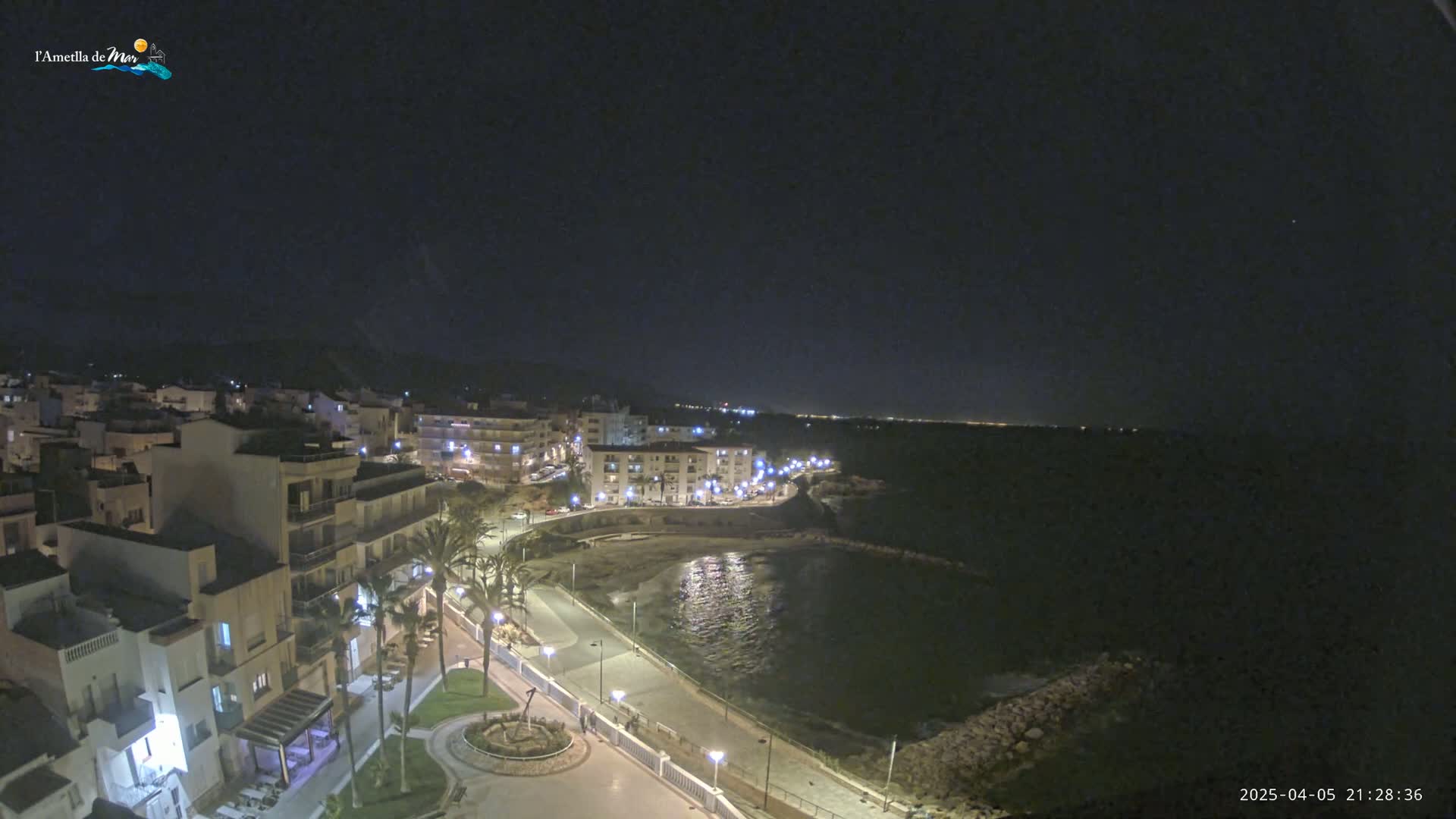 A nighttime, aerial view of a coastal town with buildings lit up along a paved waterfront path next to a dark body of water.