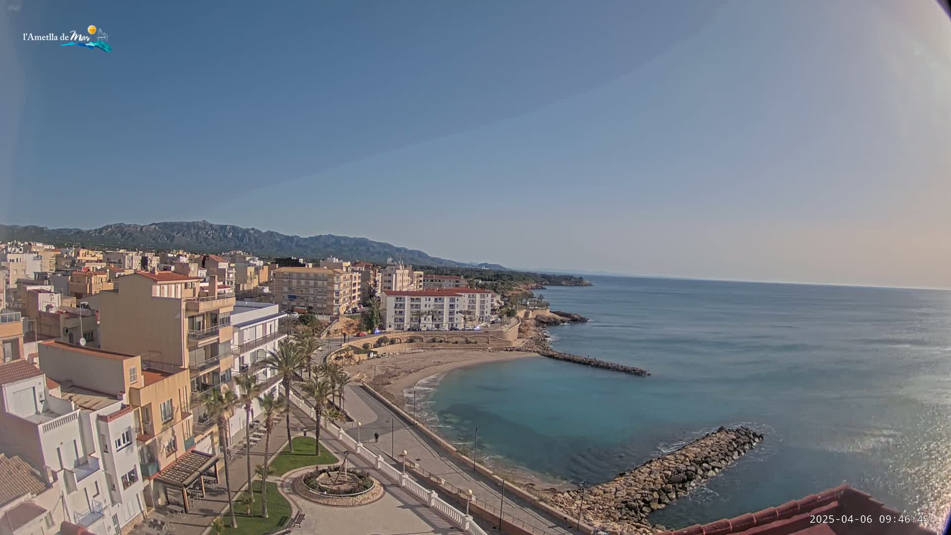 A coastal town with low-rise buildings and palm trees sits on a small, sandy cove next to a calm, blue sea under a clear, sunny sky, backed by distant mountains.