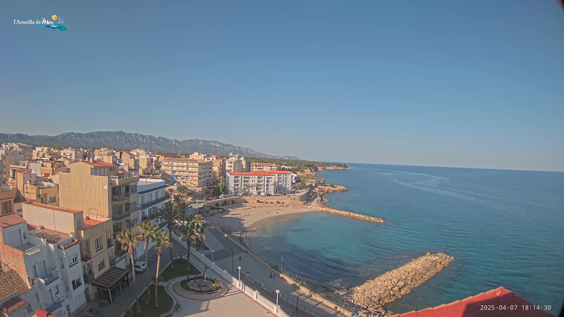A coastal town with buildings, a small sandy beach, and calm, clear blue water under a clear blue sky.