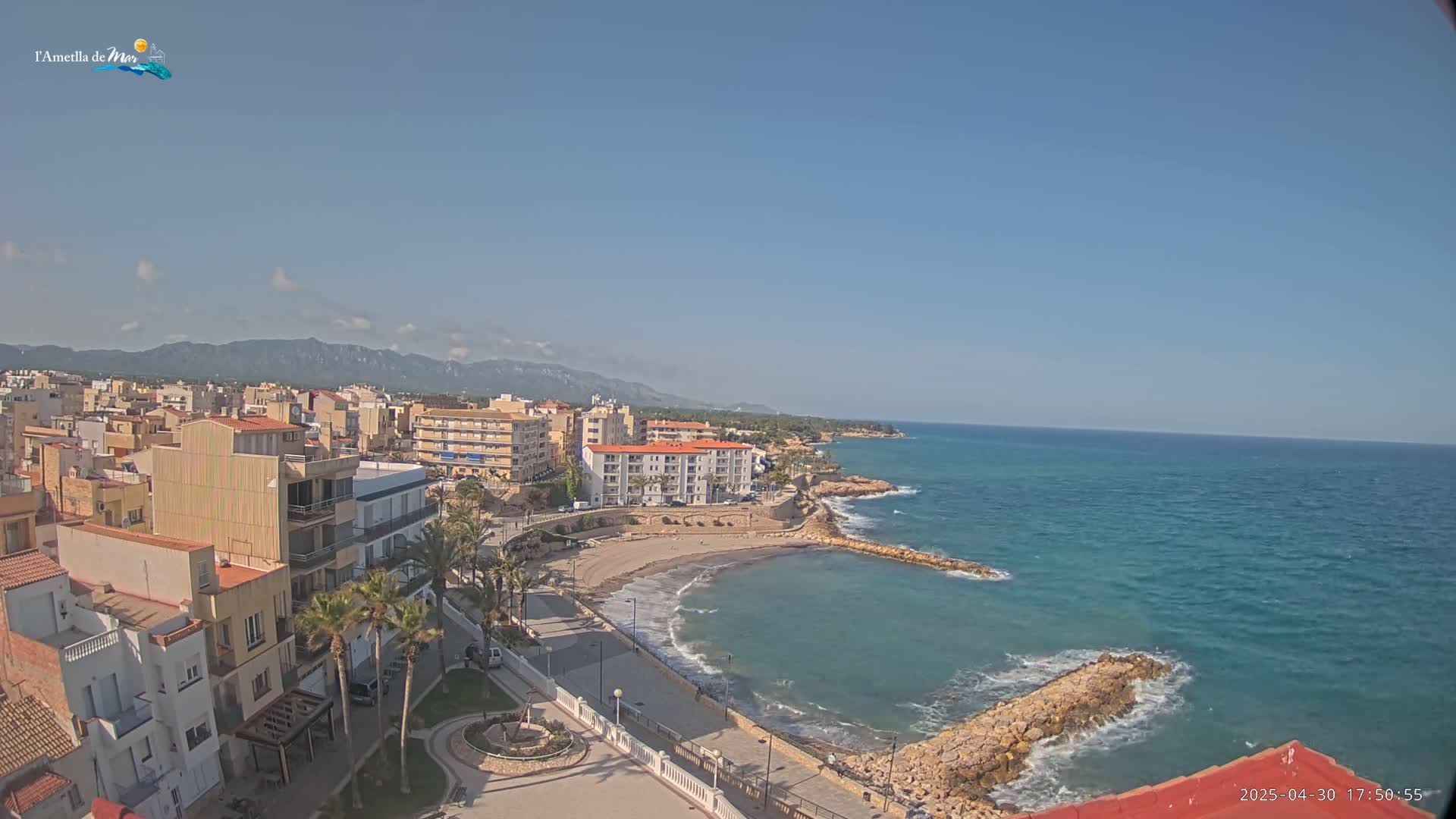 A coastal town with buildings, palm trees, and a small beach is situated against a backdrop of mountains under a clear, sunny sky.