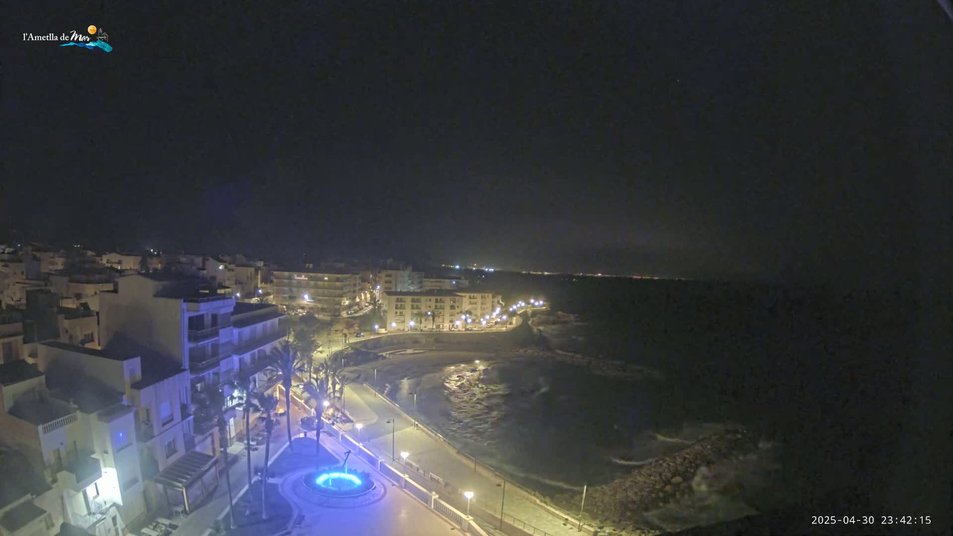 A nighttime, coastal town scene shows a crescent-shaped bay with a paved walkway, buildings with lights on, and palm trees, under a clear, dark sky.