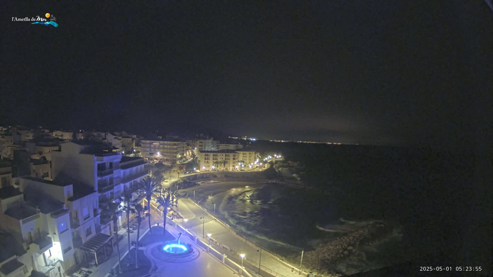 A nighttime coastal town scene shows buildings illuminated along a curving roadway that follows a dark, wavy ocean shoreline.