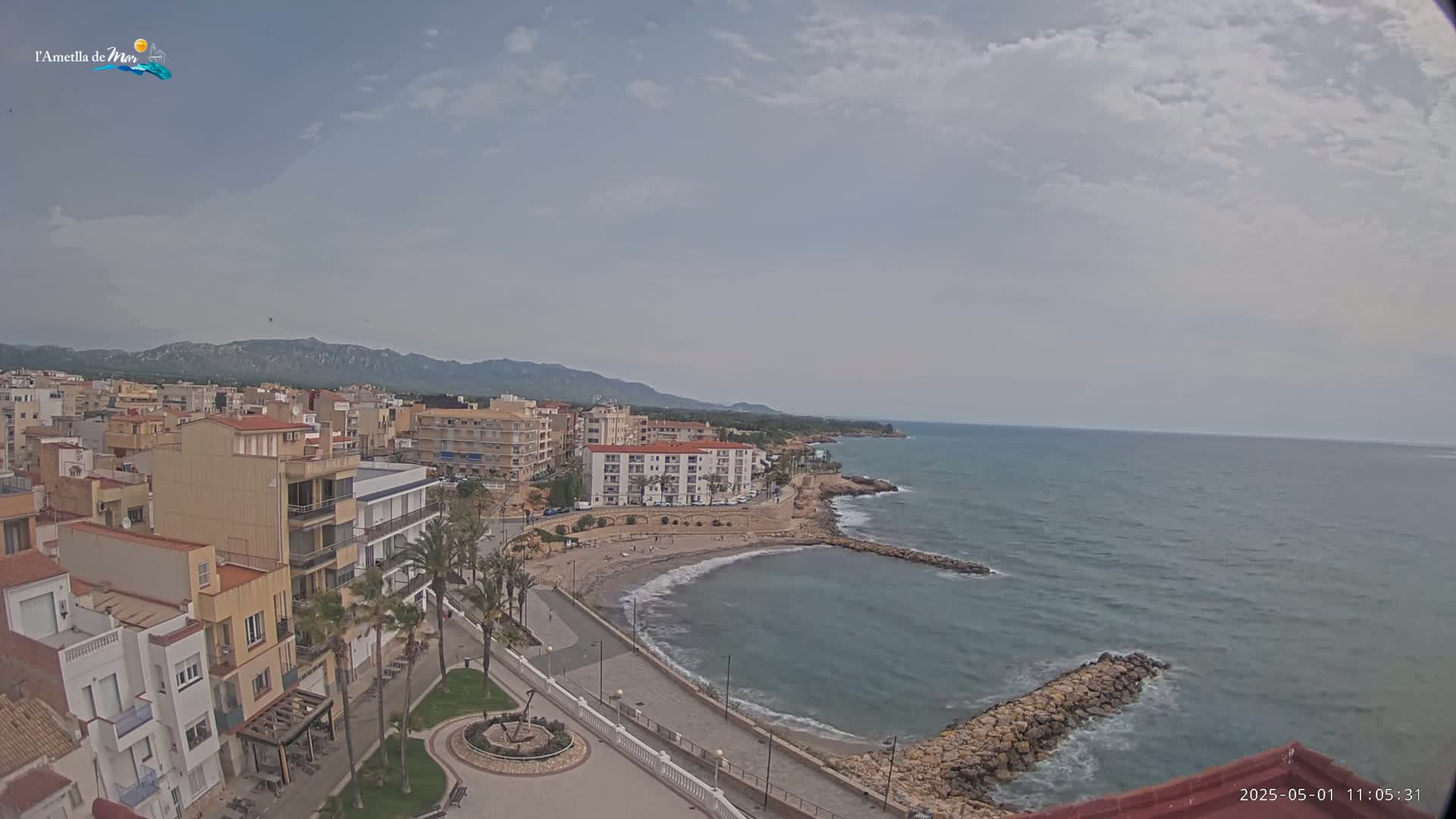 An aerial view shows a coastal town on a partly cloudy day, with buildings lining a sandy beach and calm ocean waters.
