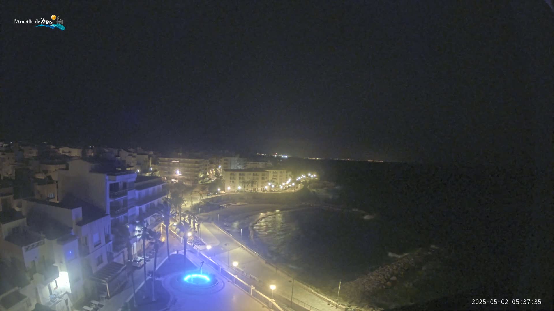 A nighttime coastal town scene shows a crescent-shaped bay with calm water, illuminated buildings along a paved waterfront, and palm trees.