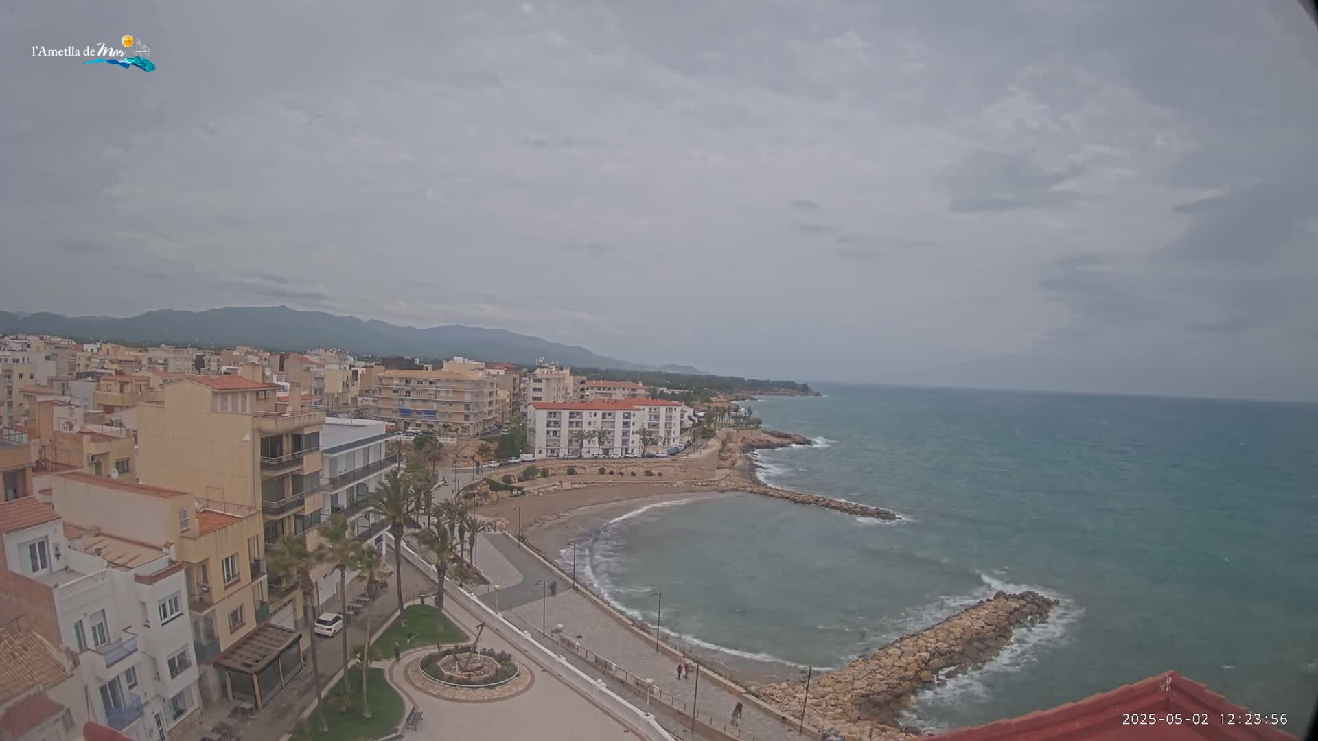 A coastal town with low-rise buildings sits under an overcast sky next to a calm sea with small waves breaking against a rocky shore.