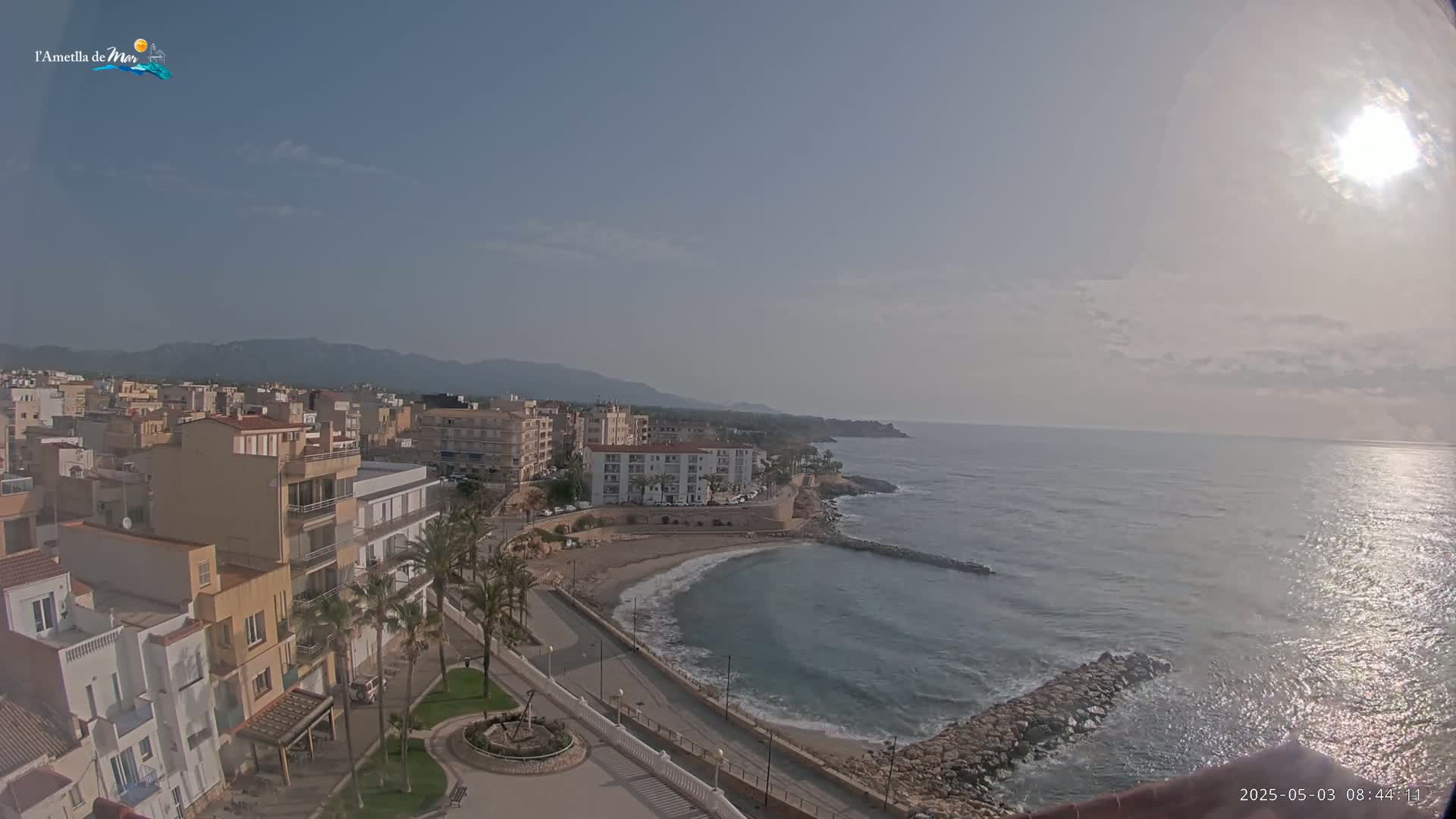 A sunny day reveals a coastal town with buildings clustered near a calm, curving bay, backed by mountains under a mostly clear sky.