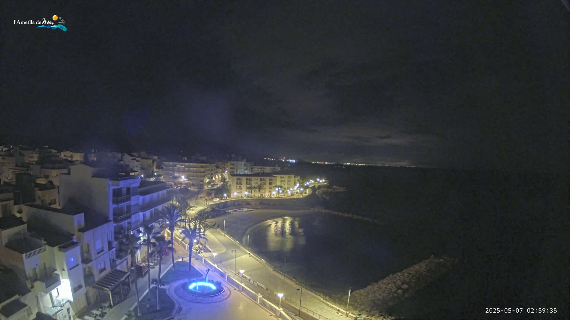 A nighttime aerial view shows a coastal town with buildings illuminated, a calm bay, and a mostly clear sky.