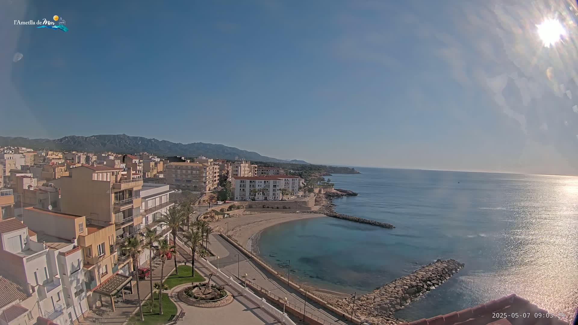 A sunny day reveals a coastal town nestled against mountains, with a calm, blue sea curving into a small sandy cove.