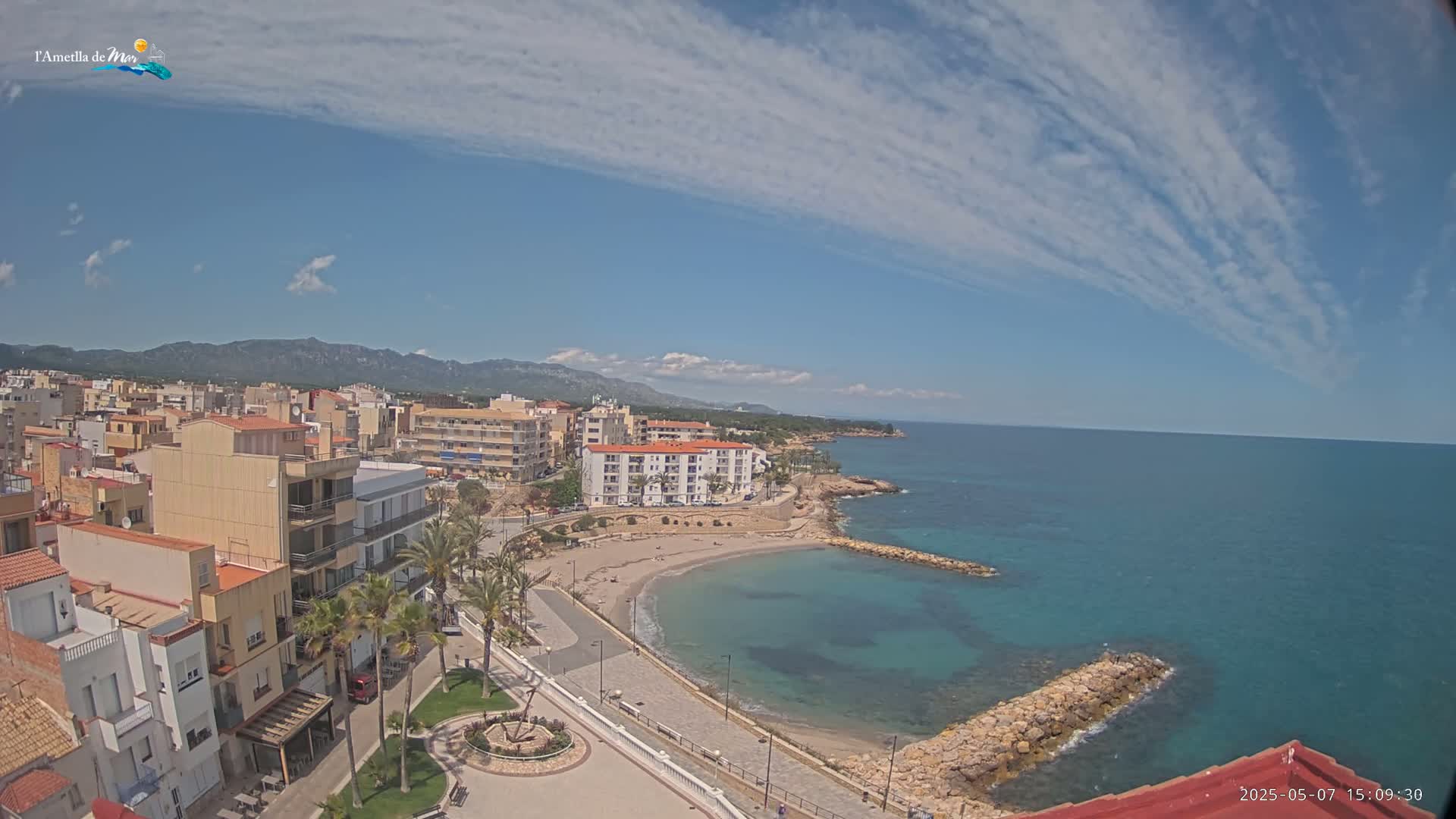 An aerial view shows a coastal town with buildings, palm trees, a small sandy beach curving into a calm teal sea under a mostly sunny sky with thin, streaky clouds.