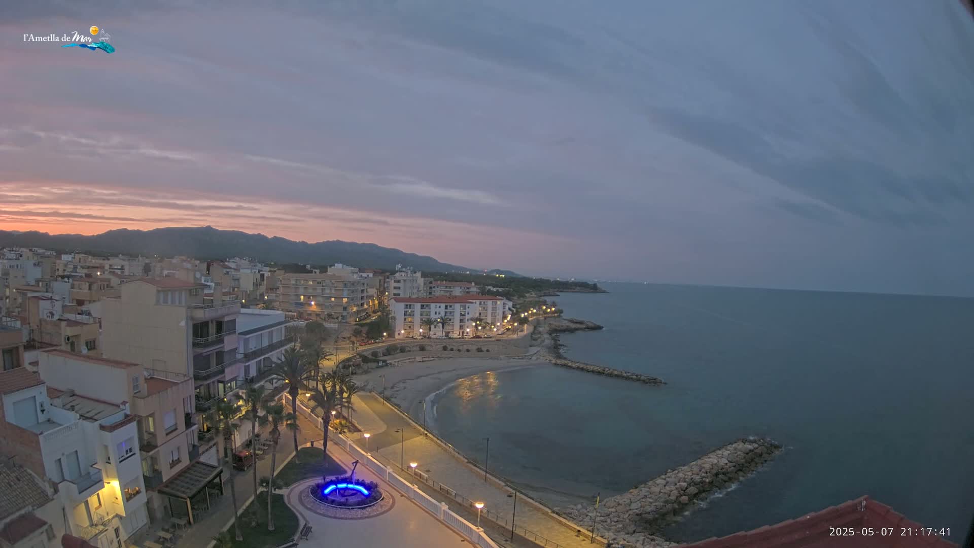 A coastal town at dusk, under a mostly cloudy sky, shows a calm sea with a small sandy beach and a promenade lined with buildings and palm trees.
