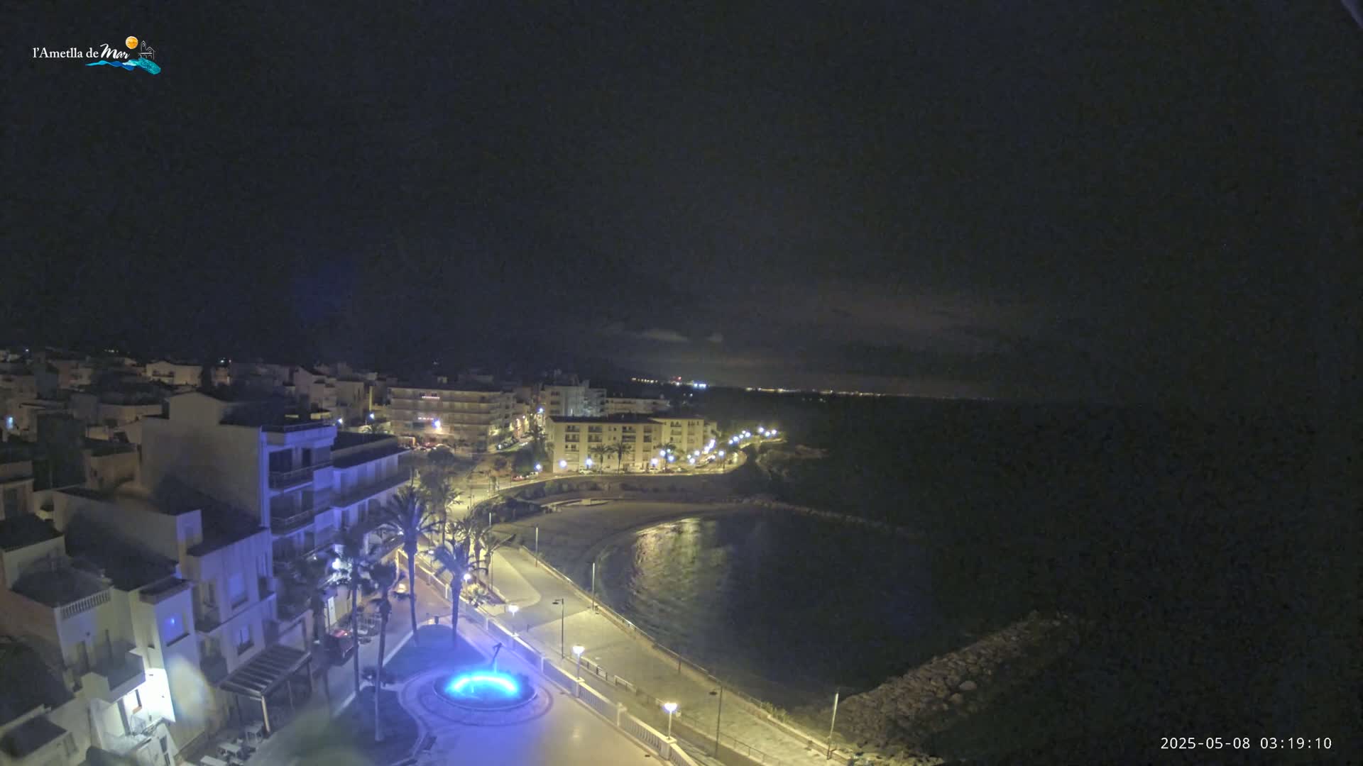 A nighttime coastal town scene shows a crescent-shaped bay with a paved walkway lined with palm trees and lit buildings, under a dark sky.