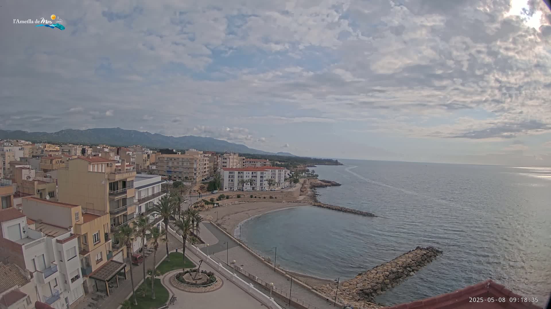 A coastal town with buildings, palm trees, and a small beach is visible under a partly cloudy sky, overlooking a calm sea with a rocky breakwater.