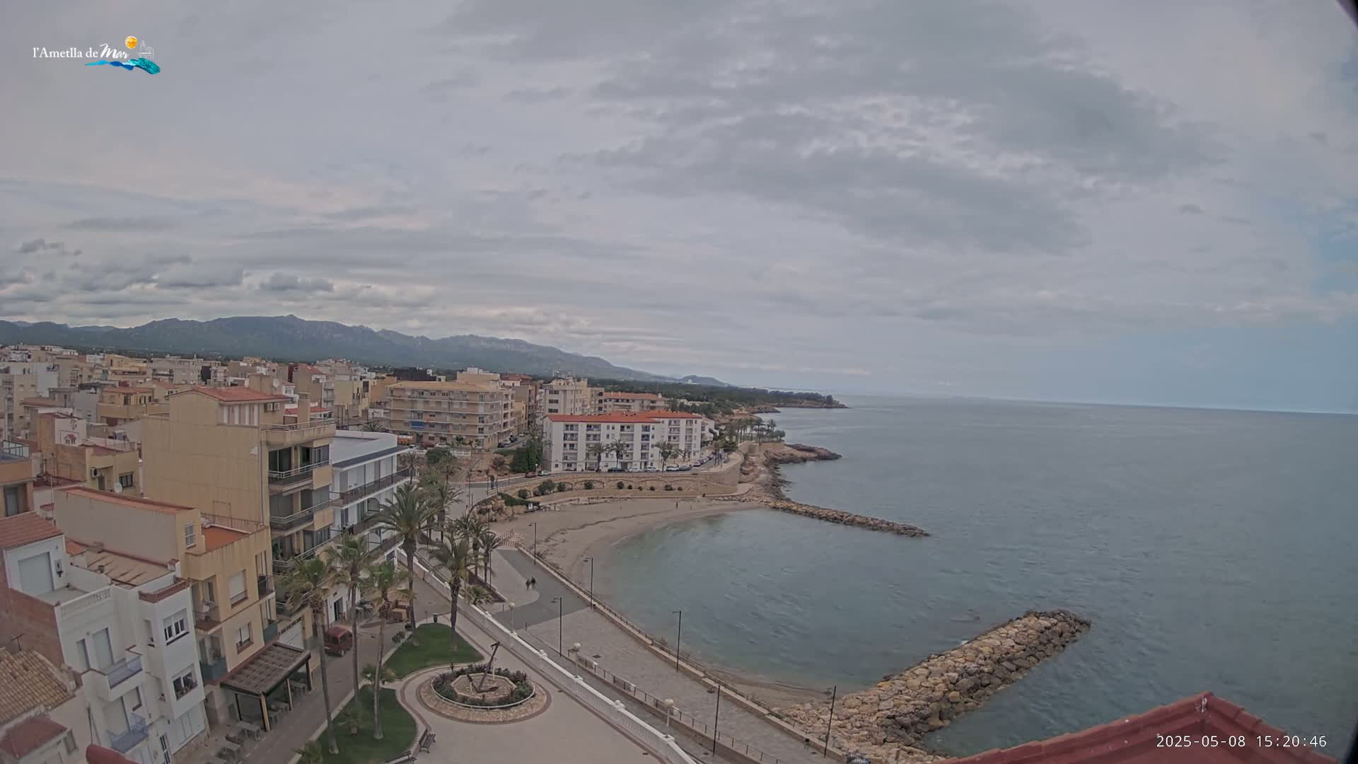 A coastal town with buildings and a small beach is visible under a cloudy sky, with calm, blue-green water and distant mountains in the background.