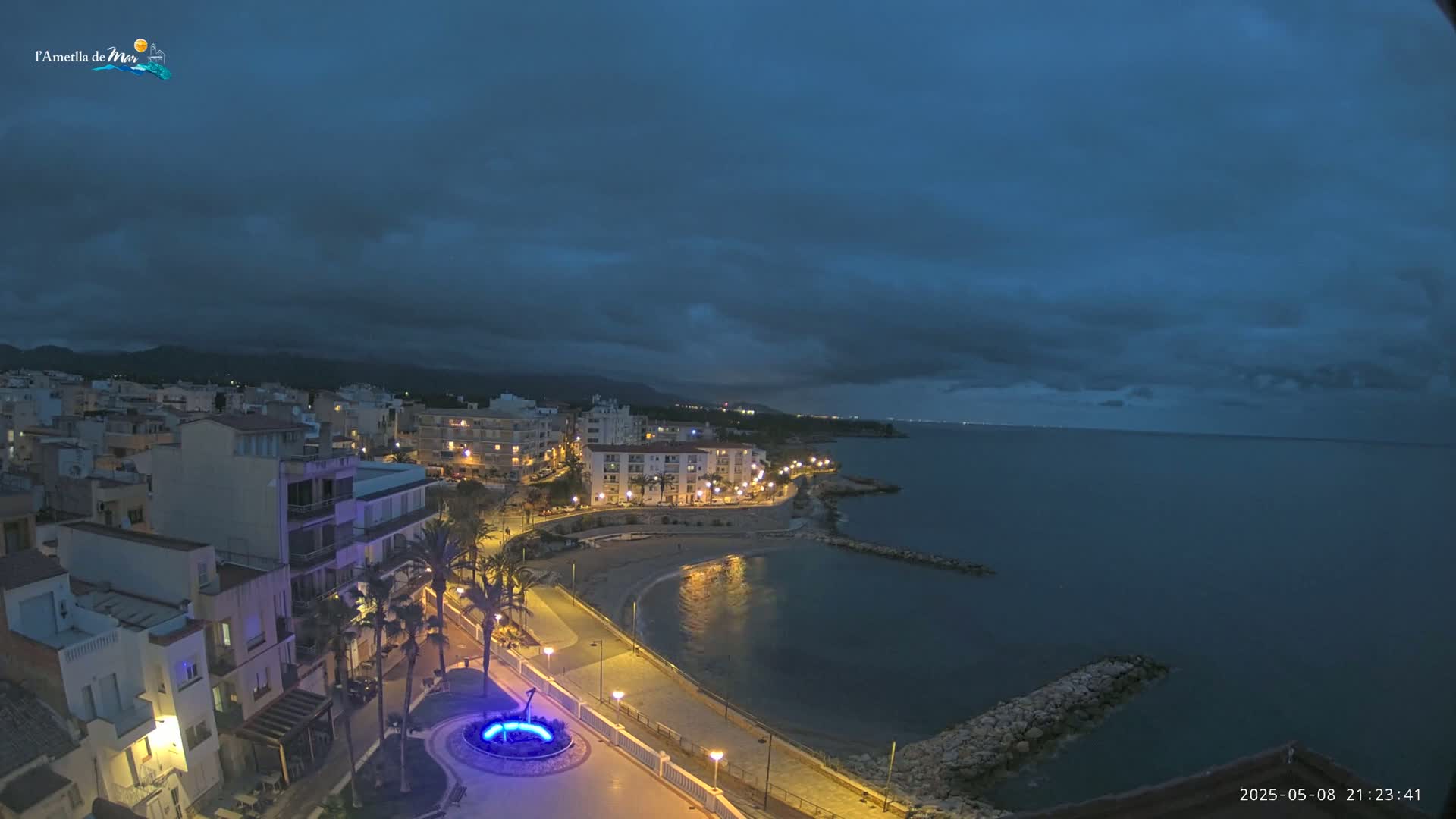 A coastal town at night under a dark, cloudy sky, with a calm sea reflecting the lights of the buildings along the shoreline.