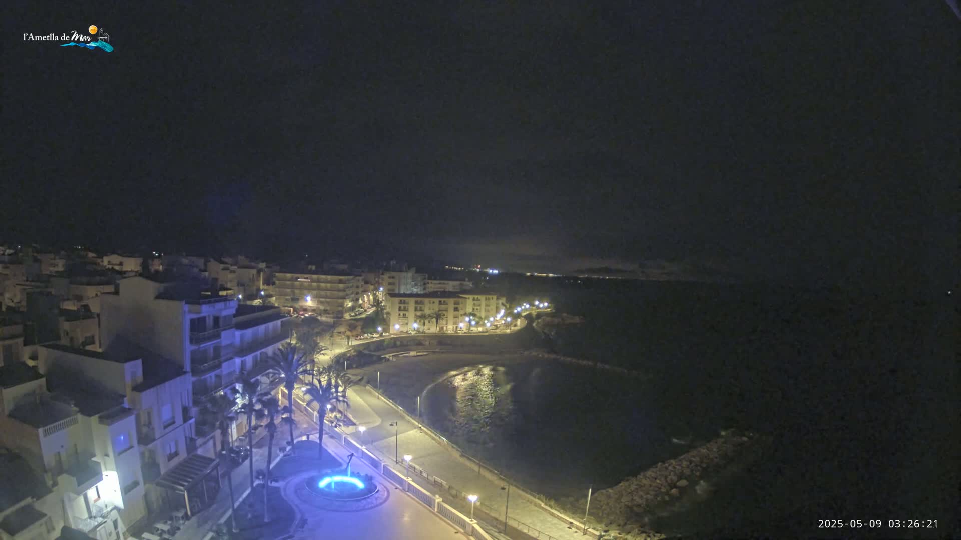 A nighttime view of a coastal town, with calm water reflecting the lights from buildings lining a curved shoreline.