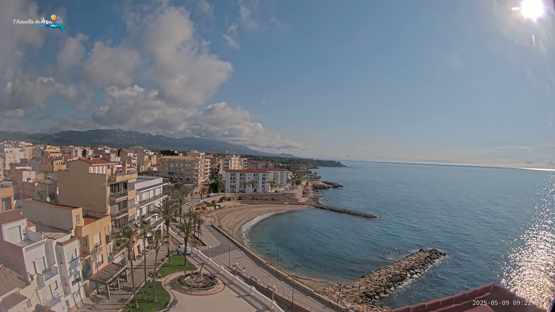 A partly cloudy day reveals a coastal town with buildings, palm trees, a small sandy beach curving into a calm blue sea, and mountains in the background.