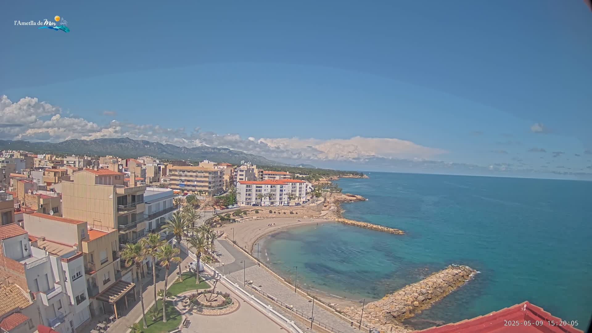 A coastal town with buildings, palm trees, a small beach, and a calm, blue sea under a mostly sunny sky with some clouds.