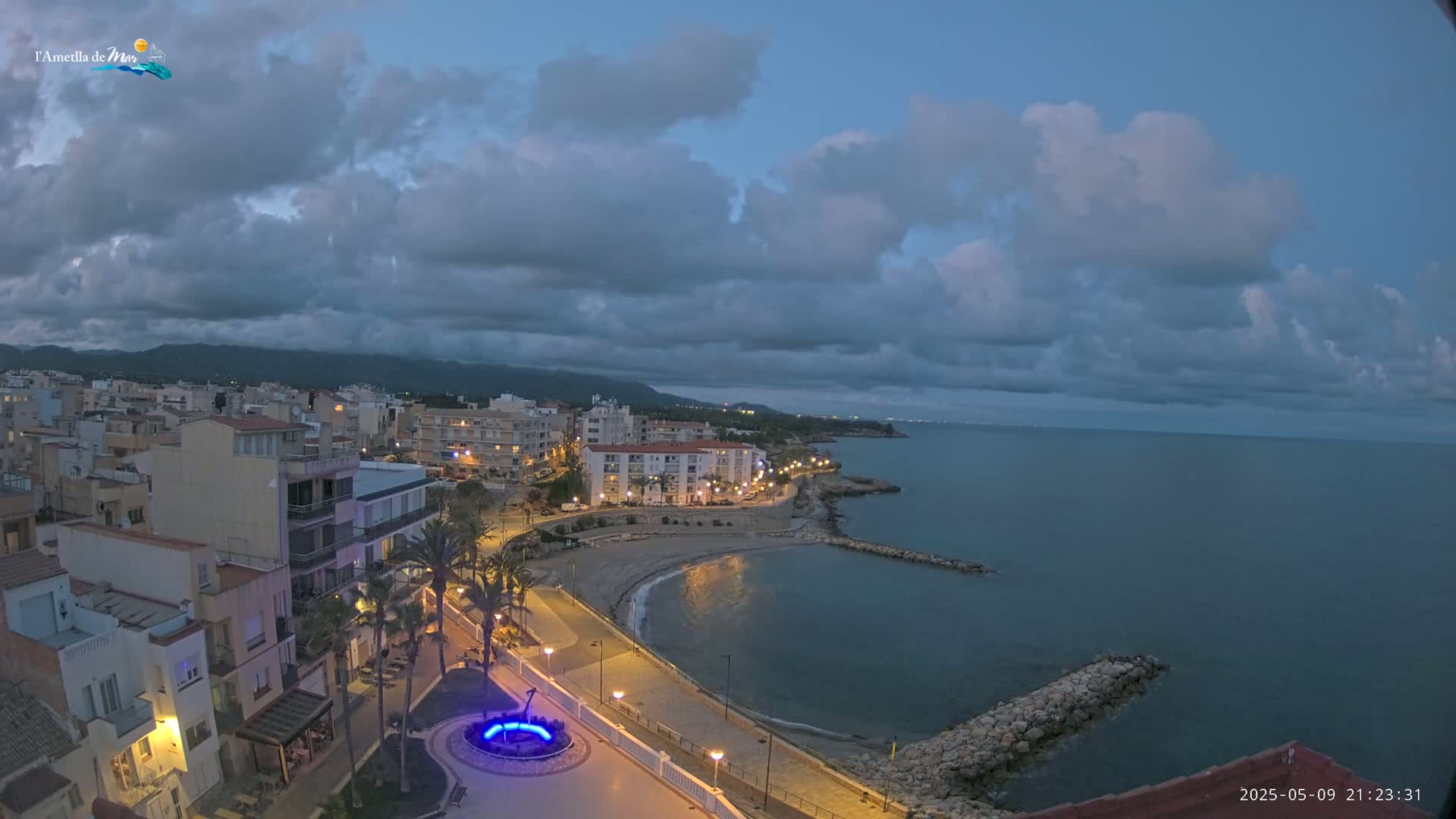 A coastal town at dusk under a cloudy sky features a crescent-shaped beach, a calm sea, and buildings illuminated along a paved walkway.