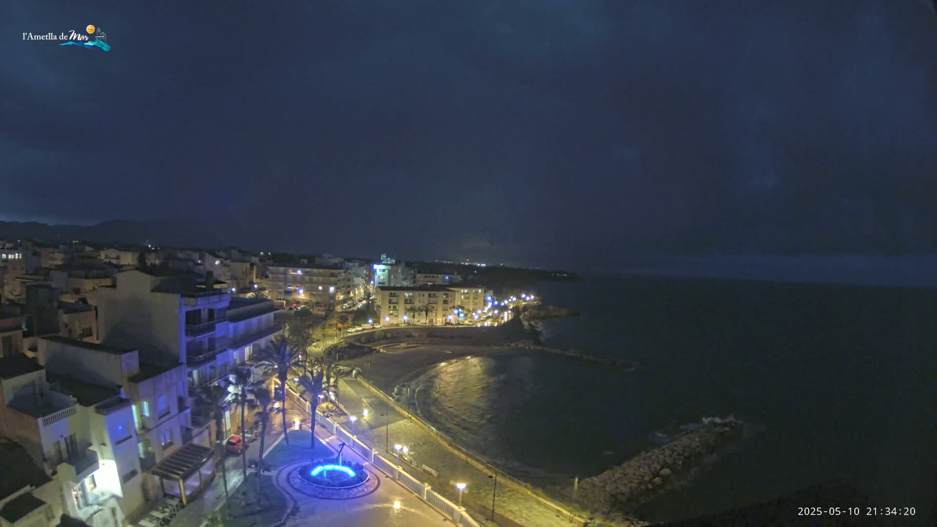 A nighttime view of a coastal town, with a calm, dark sea and illuminated buildings along a waterfront promenade.