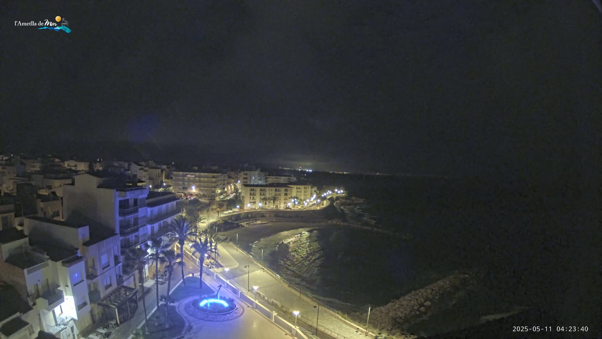A nighttime view of a coastal town with a calm sea, illuminated buildings, and palm trees lining a waterfront promenade.